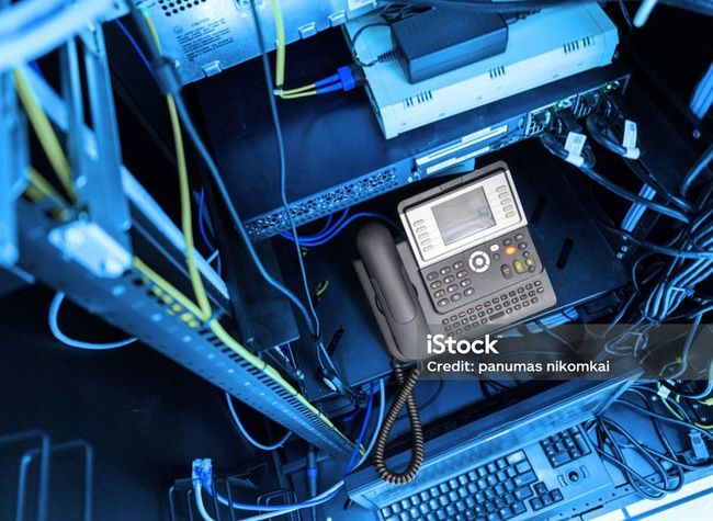 An IP phone and keyboard sit on a server rack shelf surrounded by networking cables and equipment in a dimly lit room.