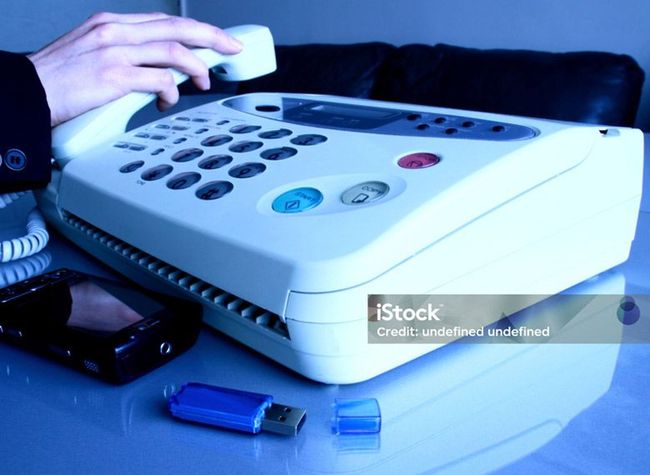 A hand picks up the receiver of a white fax machine sitting on a desk next to a black smartphone and a blue USB drive.