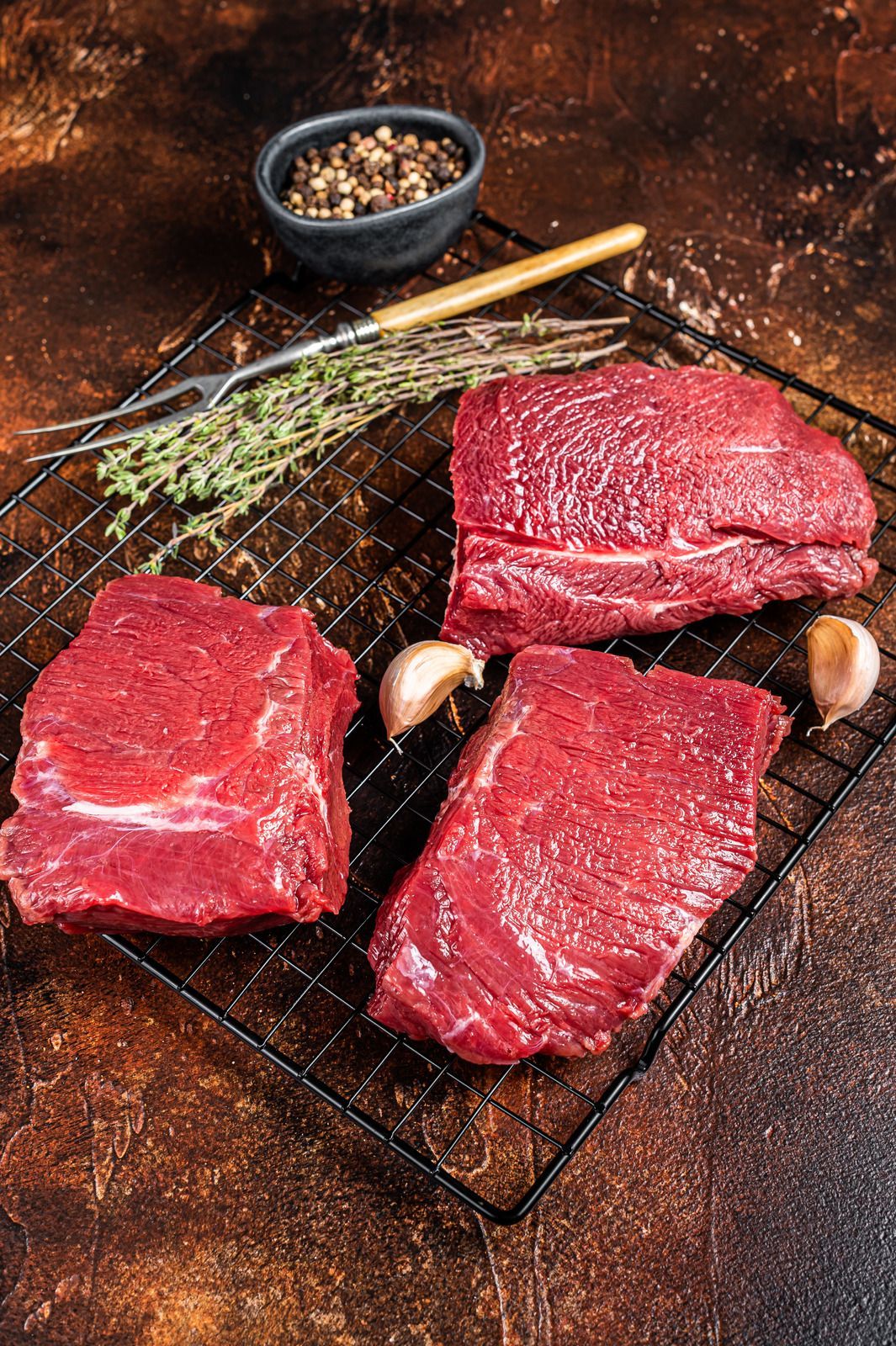 Three raw beef steaks on a metal rack with spices, herbs, and garlic cloves.
