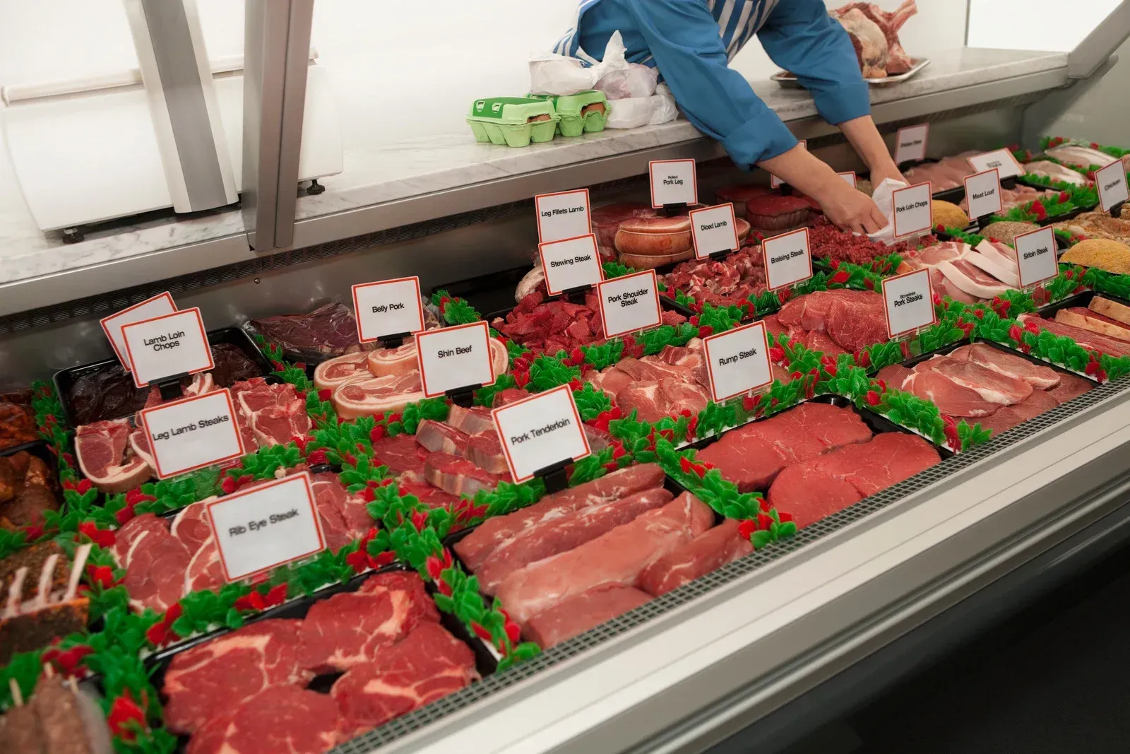 Meat display case with various cuts, labeled, and a butcher's hands arranging them.