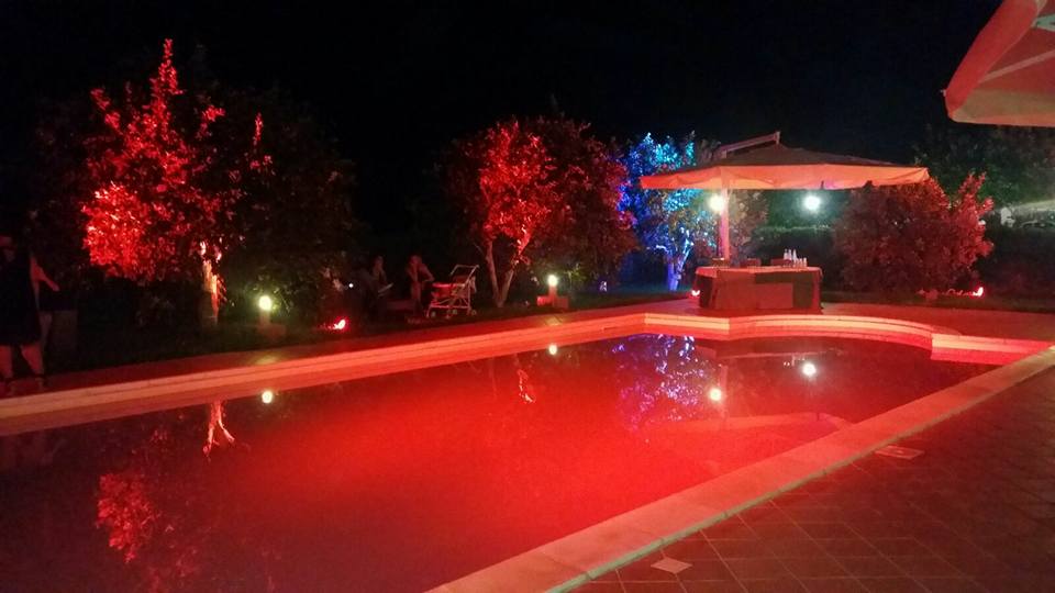 Piscina di notte, illuminata da luci rosse; gli alberi brillano di rosso e blu. Piccolo bar sullo sfondo.
