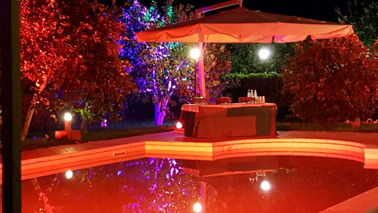 Scena notturna a bordo piscina: luci rosse e blu illuminano gli alberi, un bar sotto un ombrellone.