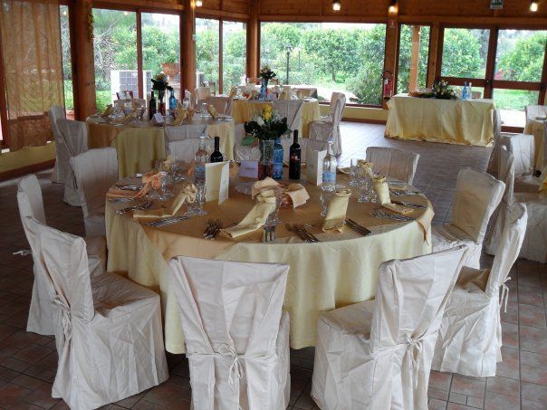 Tavoli apparecchiati per una festa in un locale con grandi finestre, tovaglie e coprisedie beige.