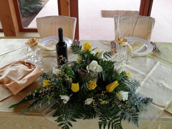 Apparecchiatura della tavola con centrotavola floreale, bottiglia di vino e segnaposto in una stanza piena di luce.