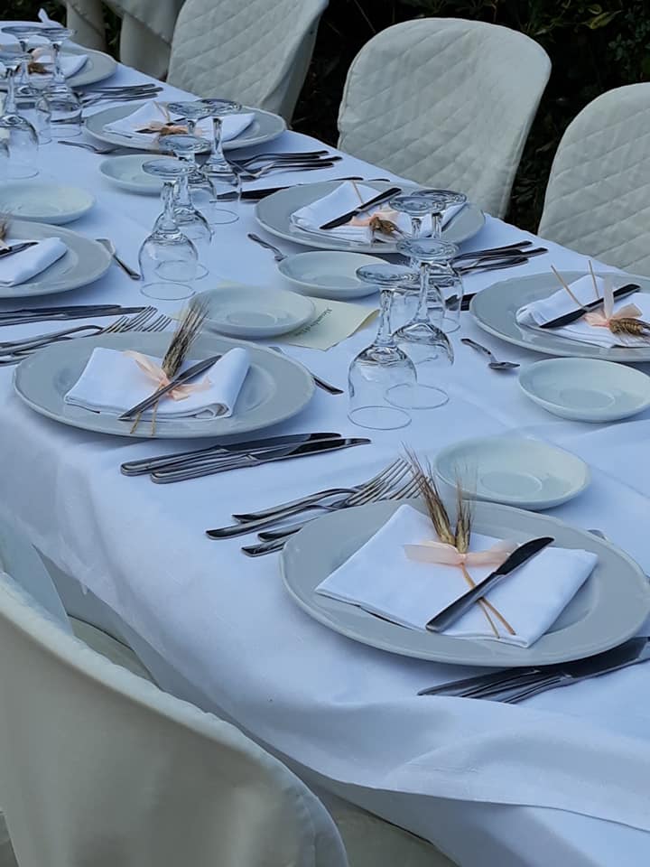 Tavolo apparecchiato per un evento formale all'aperto con tovaglia bianca, piatti, posate e bicchieri da vino.