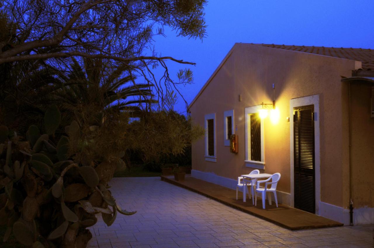 Esterno della casa al tramonto con finestre illuminate, tavolo e sedie sul patio. Palma e cielo azzurro.