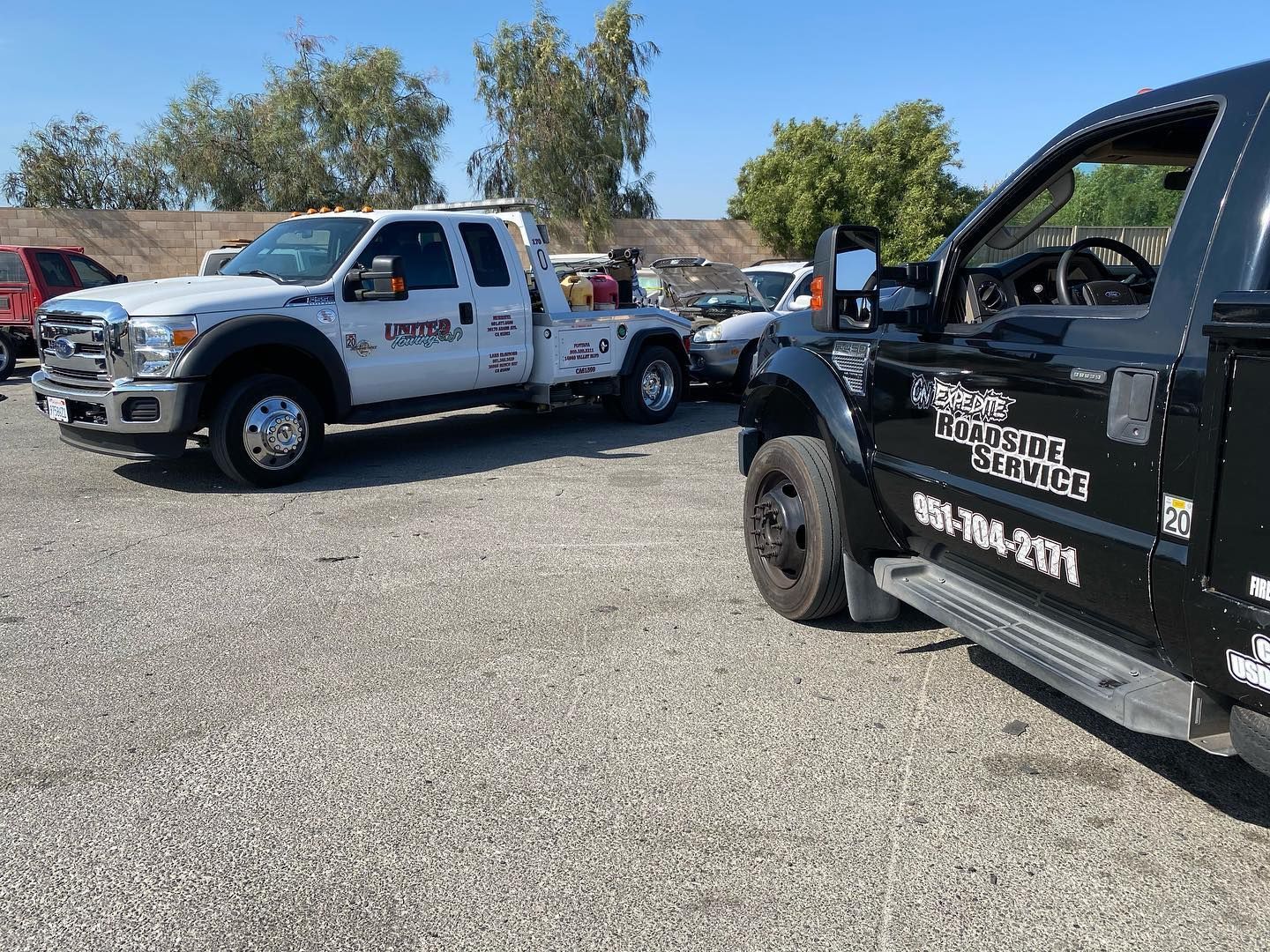 Two tow trucks parked on gravel. One white, one black. Sunny outdoor setting.