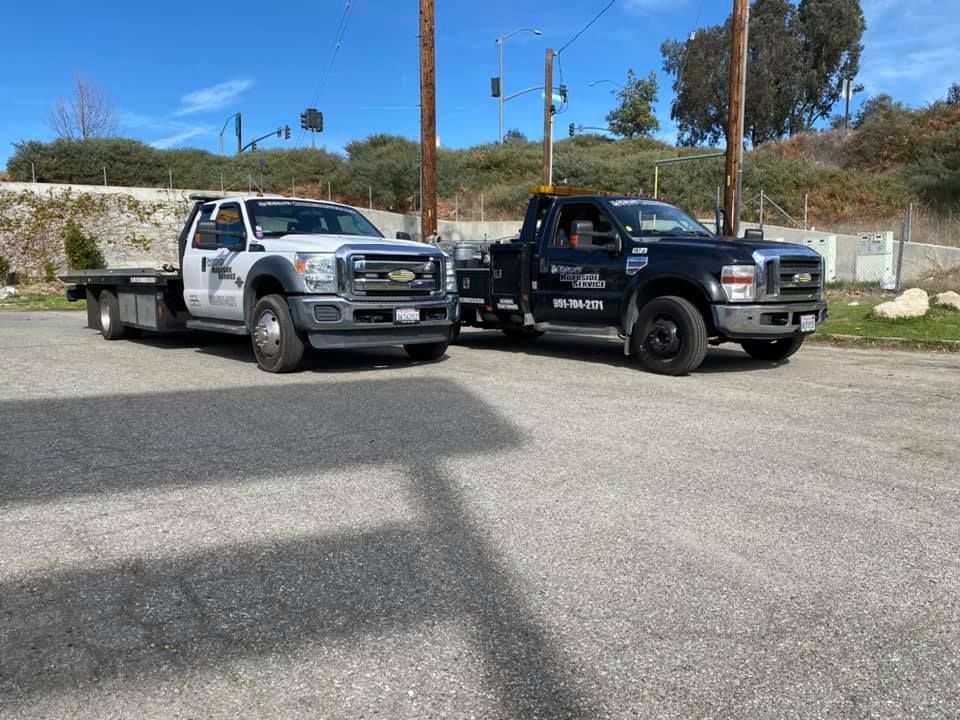 Two tow trucks parked side-by-side on a gravel lot; one white, one black.