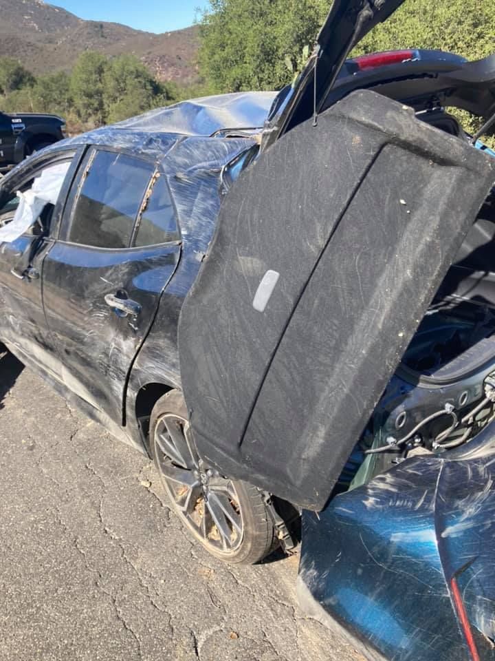 Damaged black car involved in a collision; rear hatch open, roof caved in, on a road.
