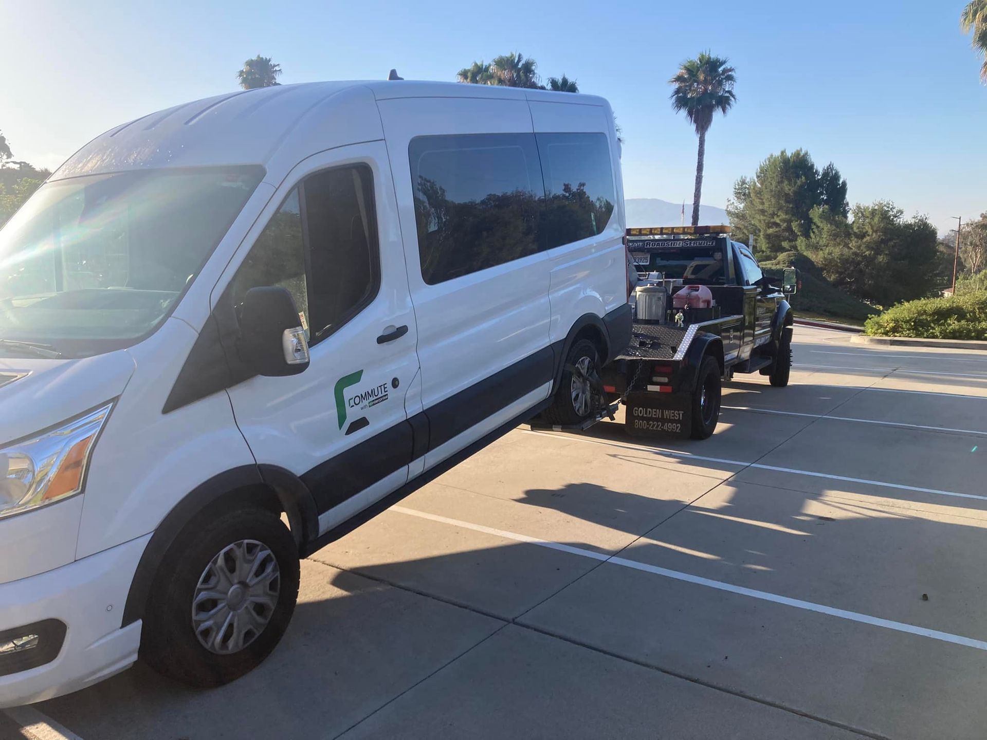 White van with logo towing a black trailer, parked on a paved surface in an outdoor setting.