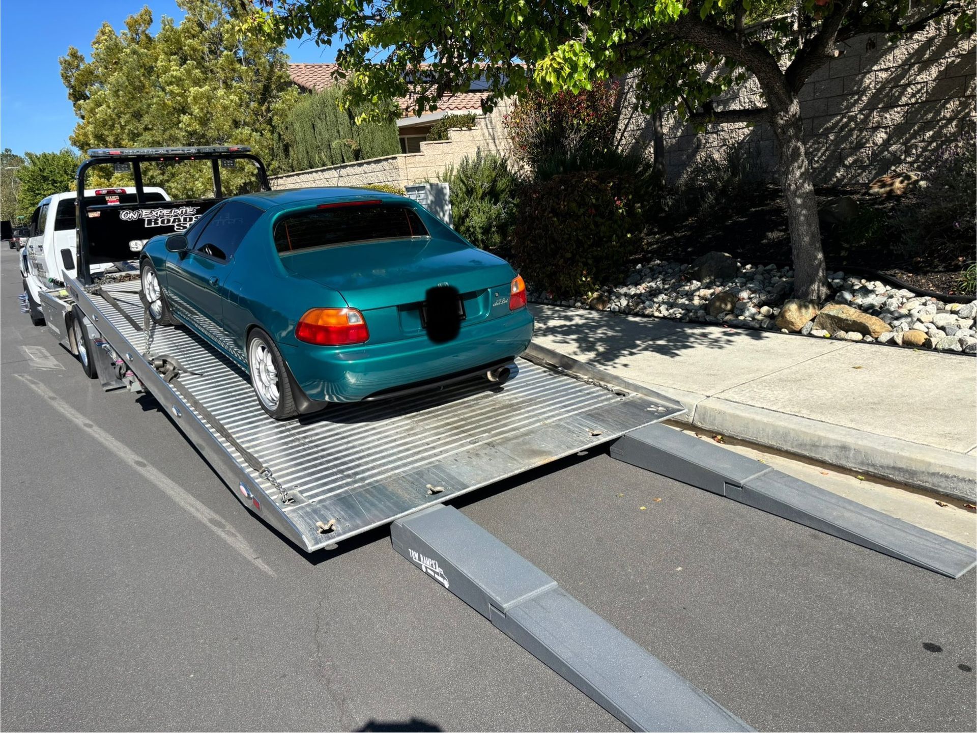 A teal Honda Del Sol is towed on a flatbed truck on a sunny street.