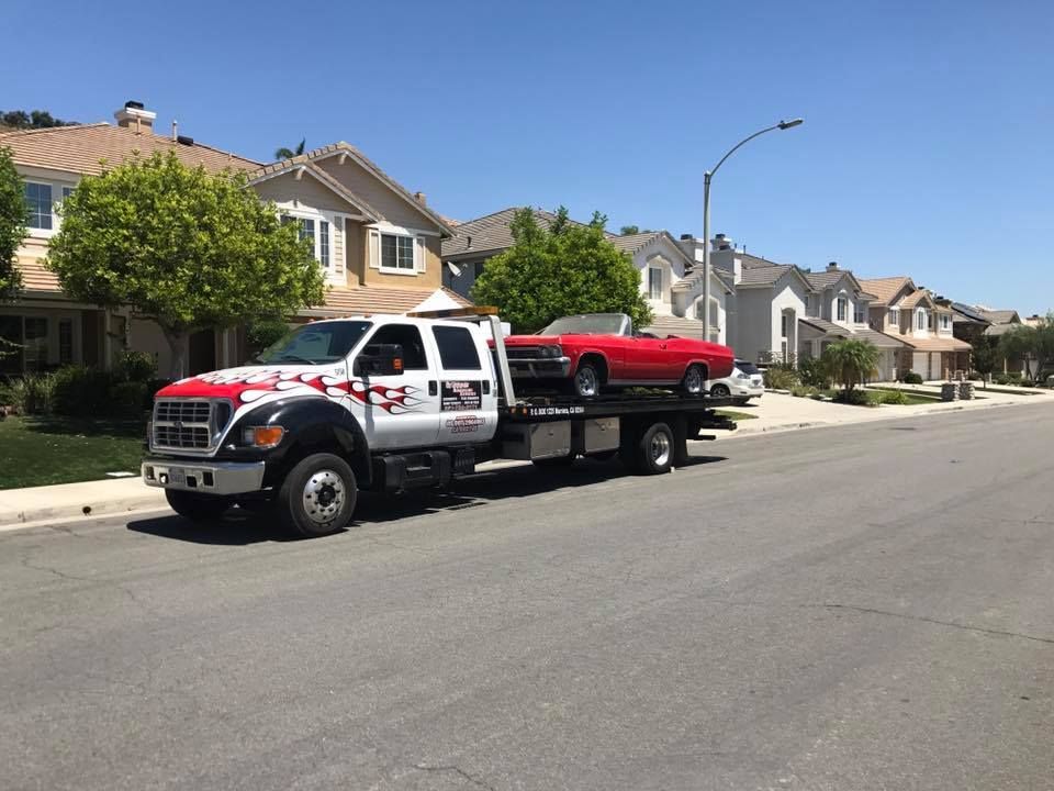 Tow truck hauling a red classic convertible on a residential street under a bright blue sky.
