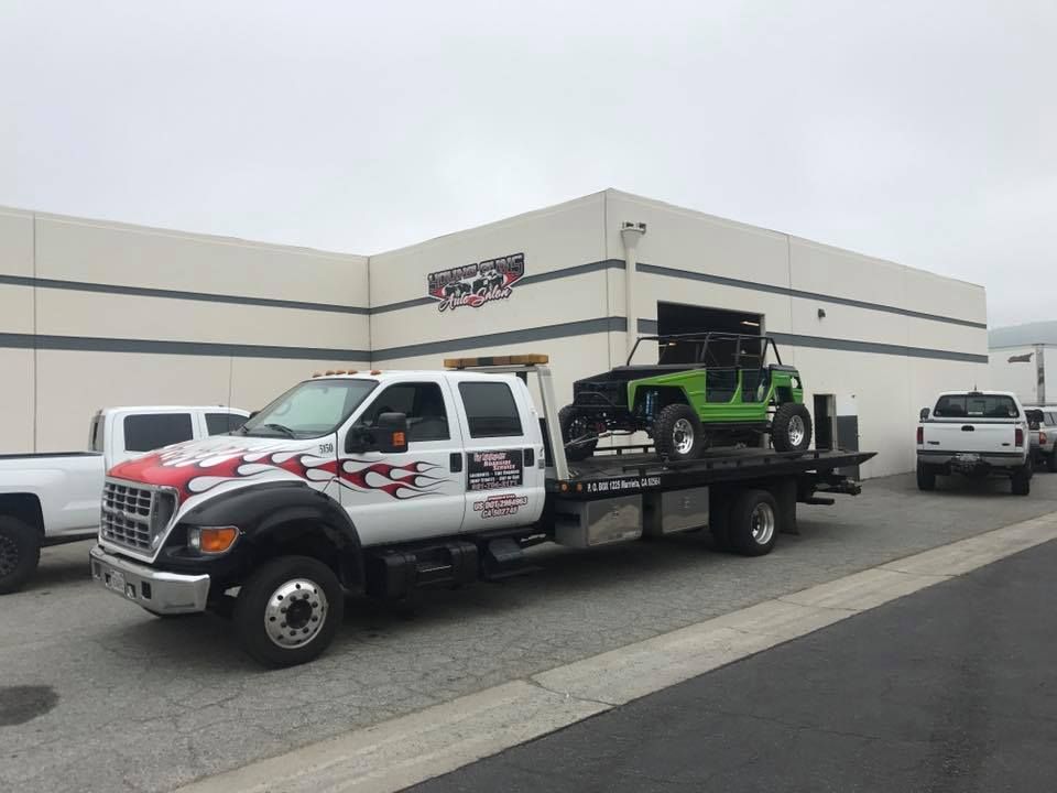 Tow truck with a green off-road vehicle on its flatbed, parked in front of a building on a cloudy day.