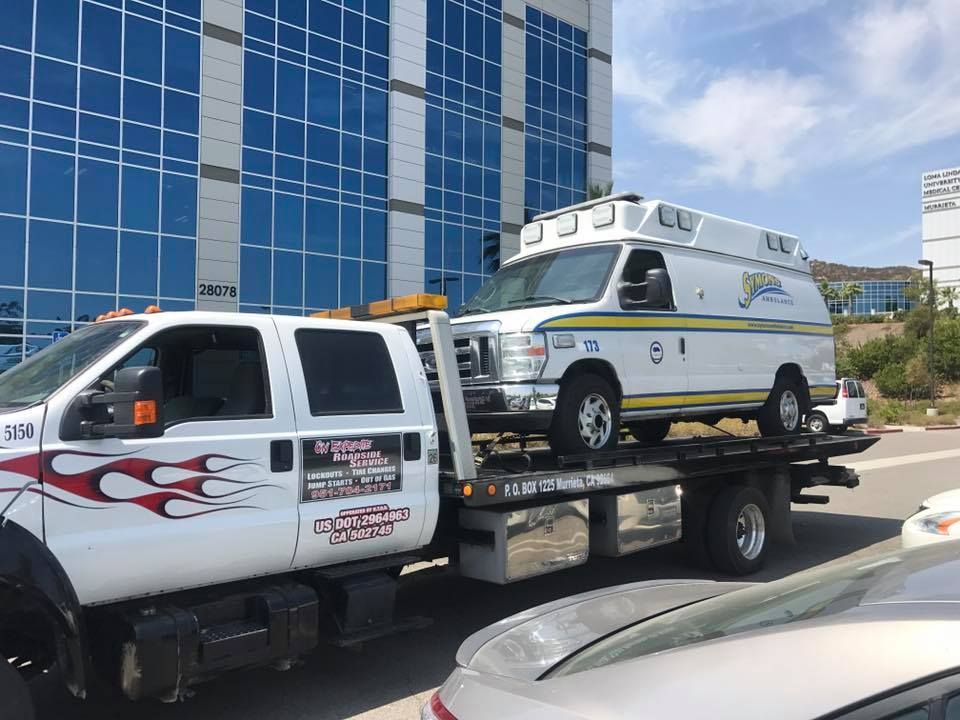 A white tow truck hauling a white van with yellow stripes near a glass-walled building.
