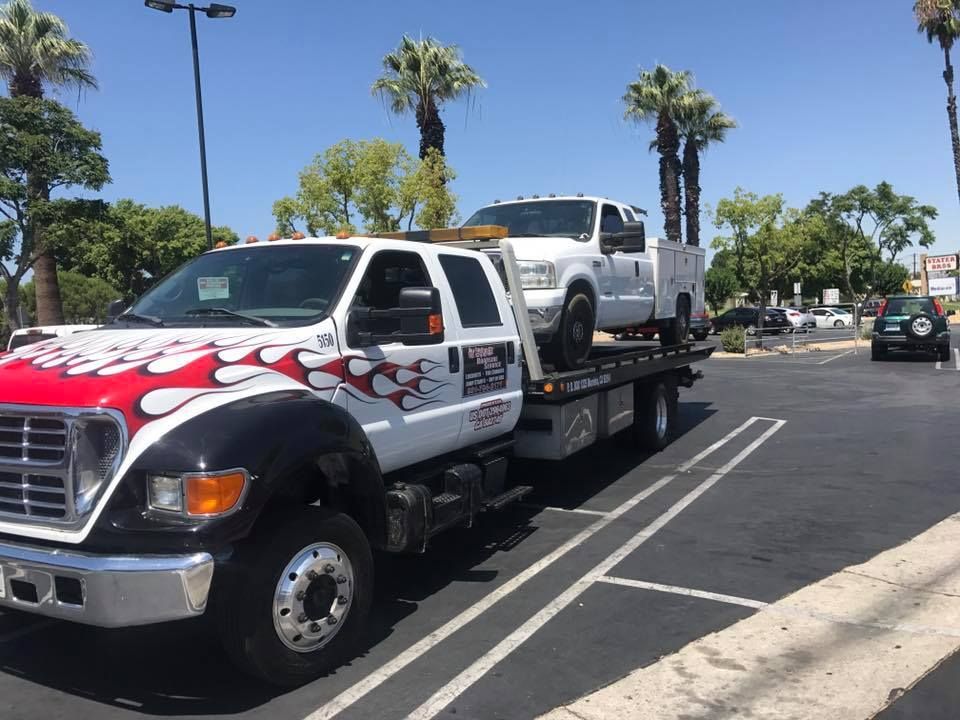 A white tow truck carrying a white pickup truck in a parking lot on a sunny day.