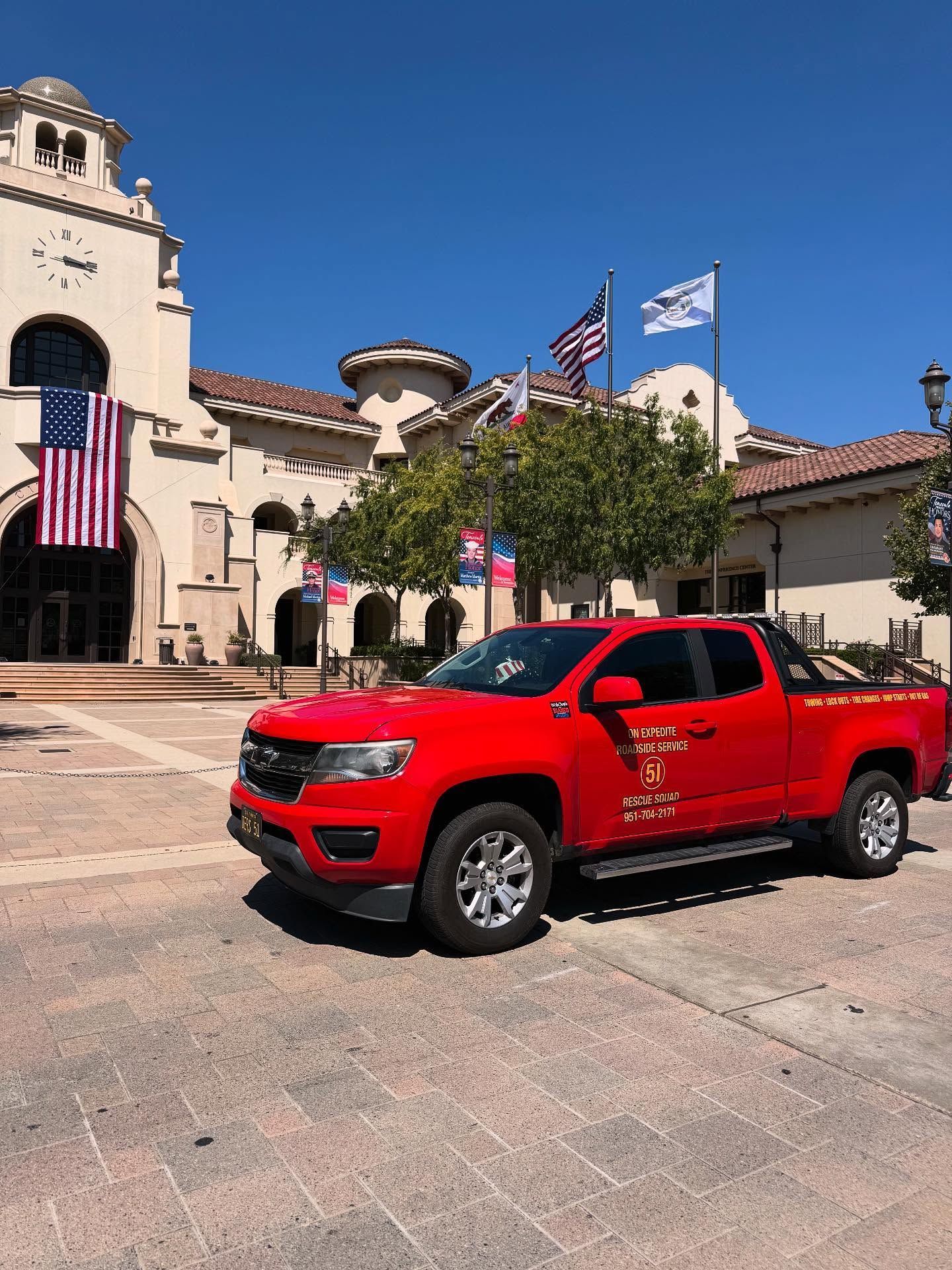 Red pickup truck parked in front of a light-colored building with American flags; sunny day.
