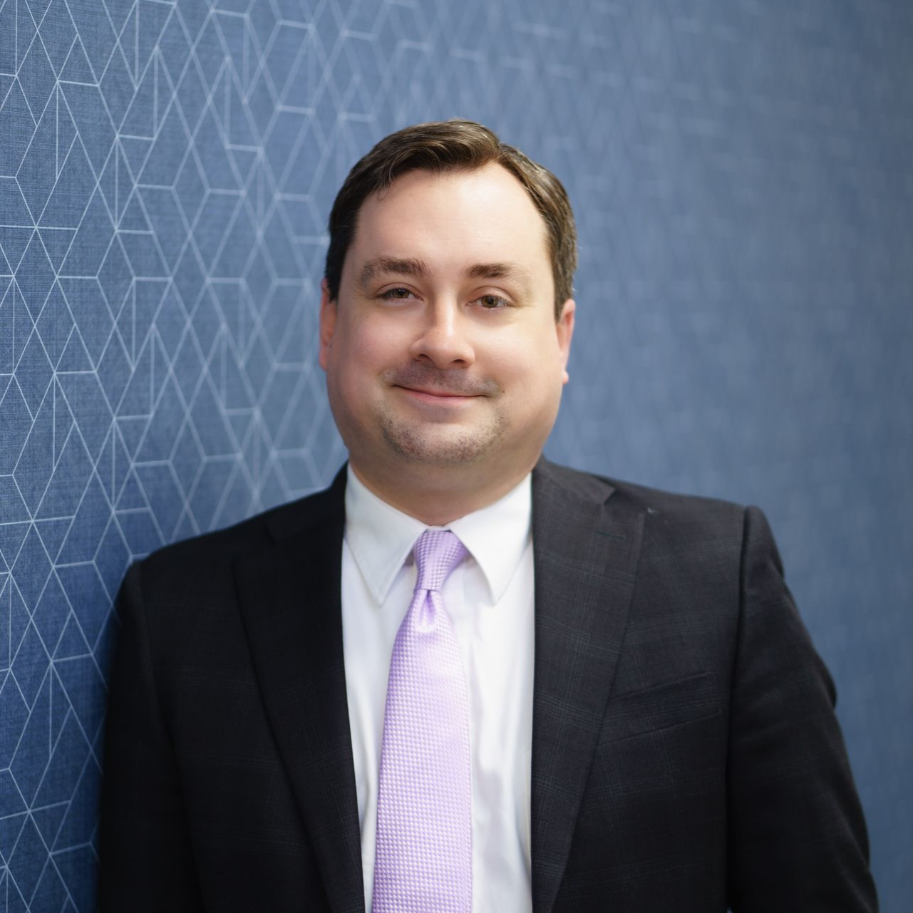 Man (Ben Yelverton) in a dark suit and light purple tie against a blue geometric background