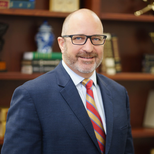 Bald man in glasses (Joel Shields) wearing a navy suit and striped tie, smiling in an office setting