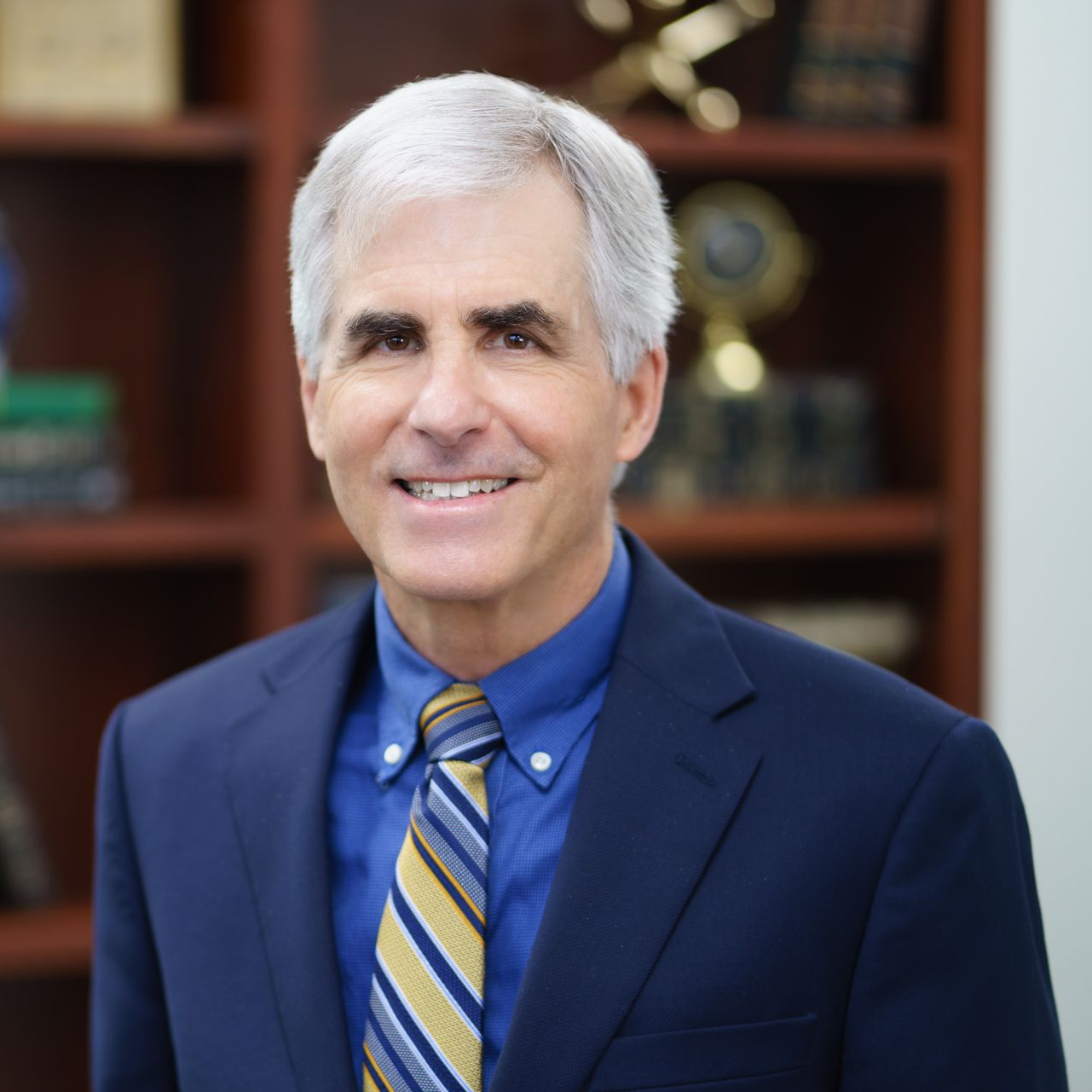 Man (Mike Scanes) in blue suit and striped tie smiling in front of a bookshelf background
