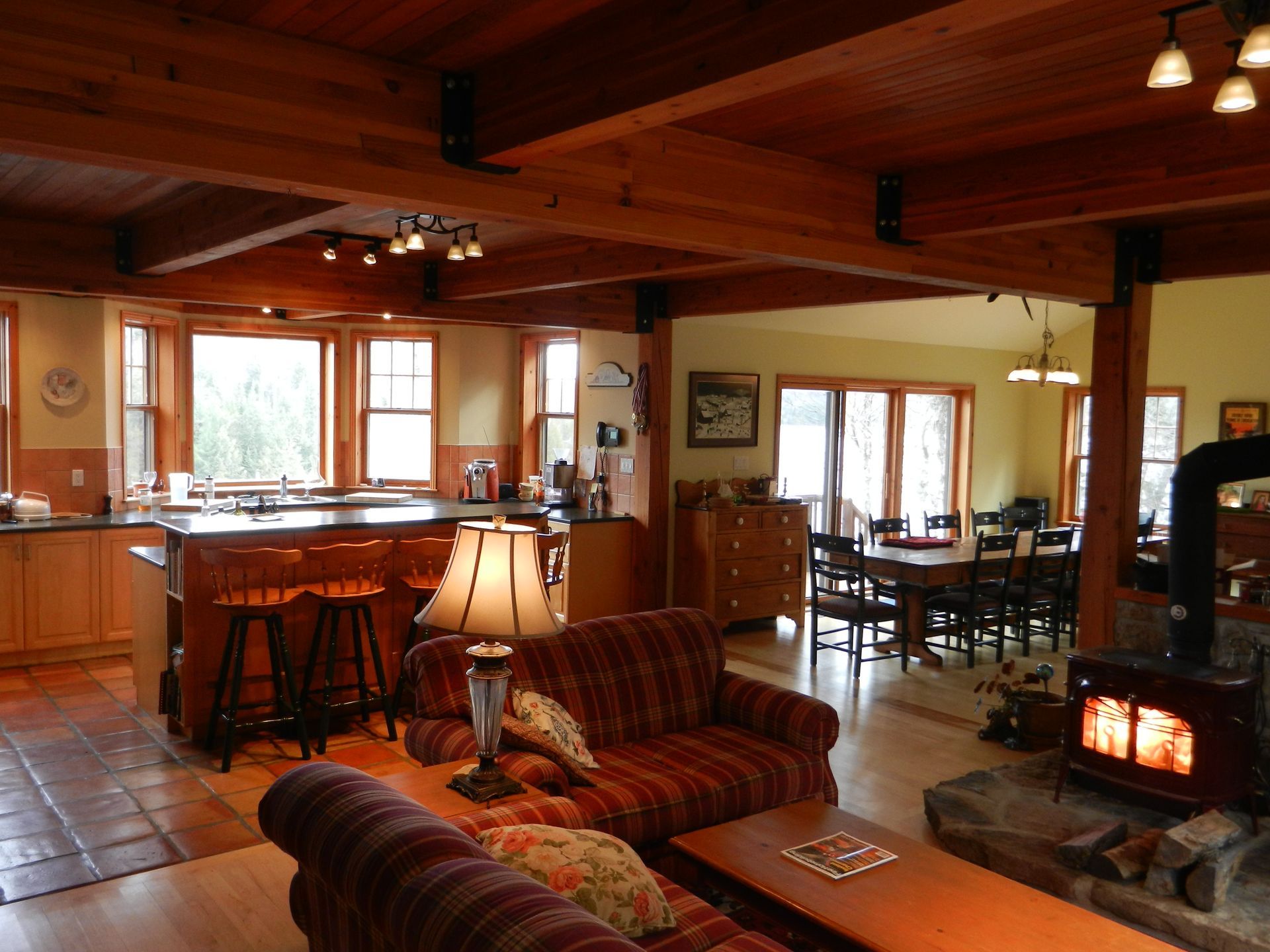 Open-plan living space with kitchen, living room, and dining area. Exposed wooden beams, cozy fireplace, and warm color scheme.