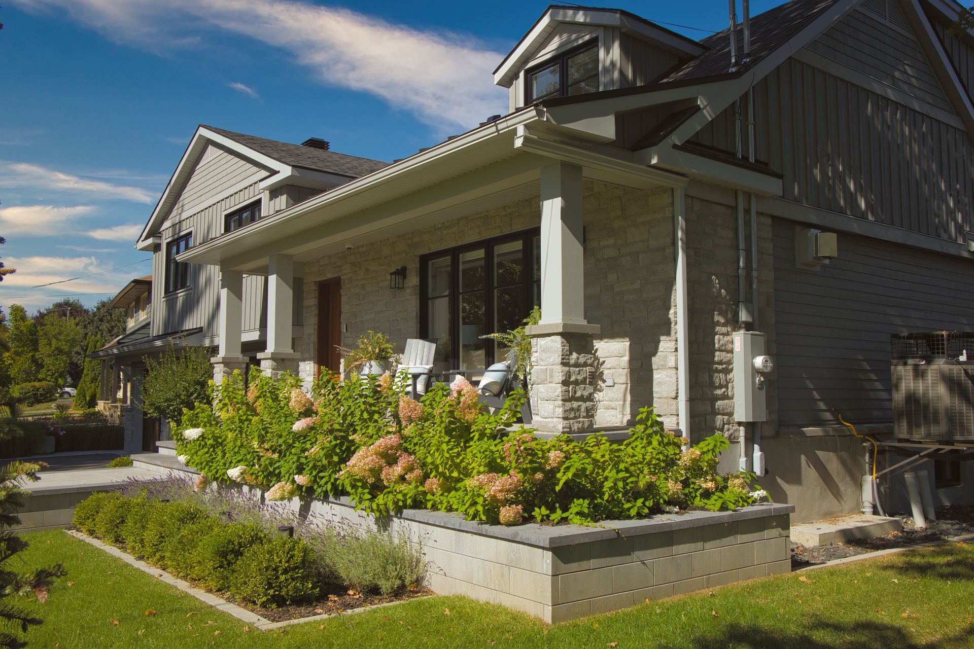 House exterior with porch, flower beds, and gray siding on a sunny day.