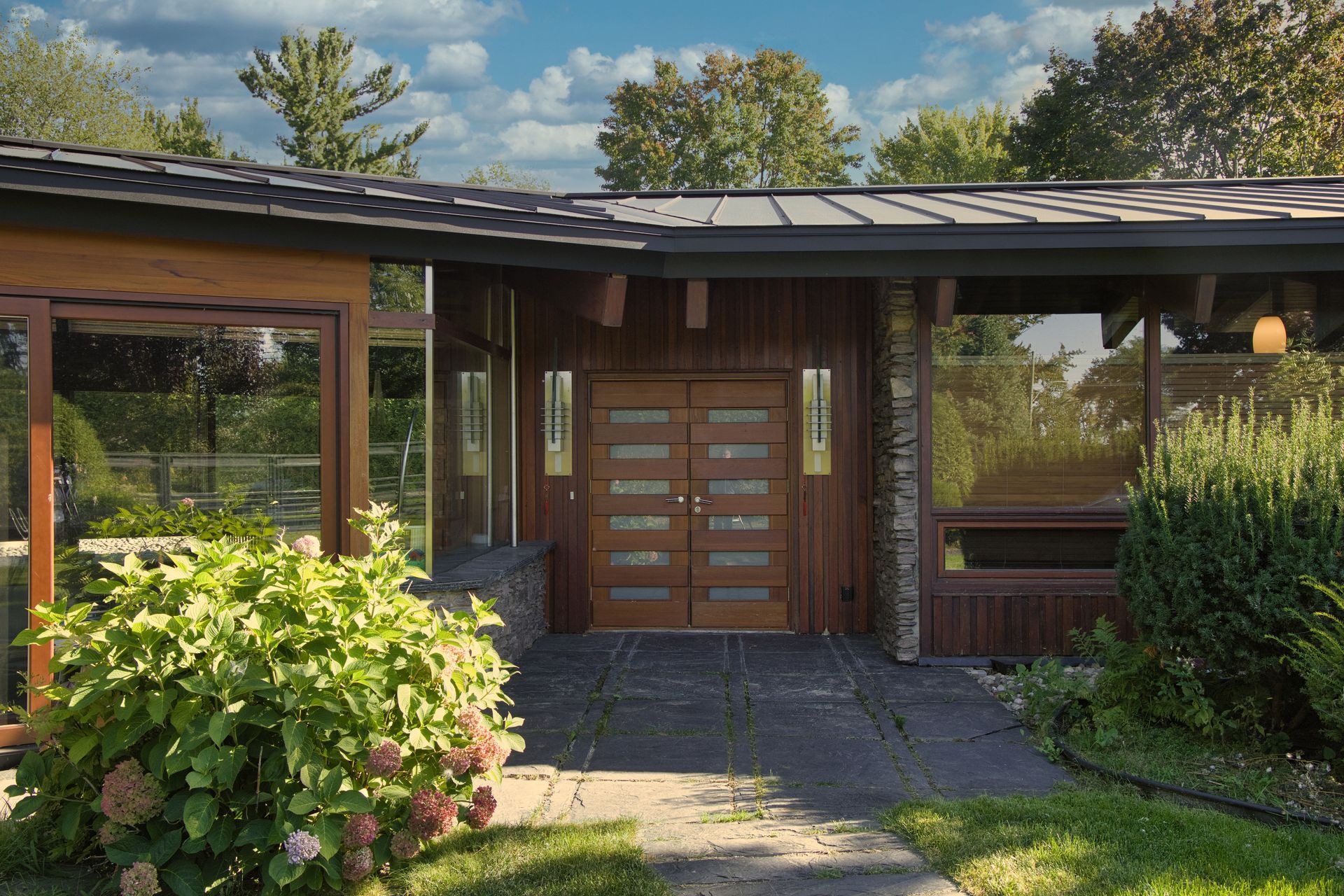 Brown home with large glass windows and double doors, green bushes in front.