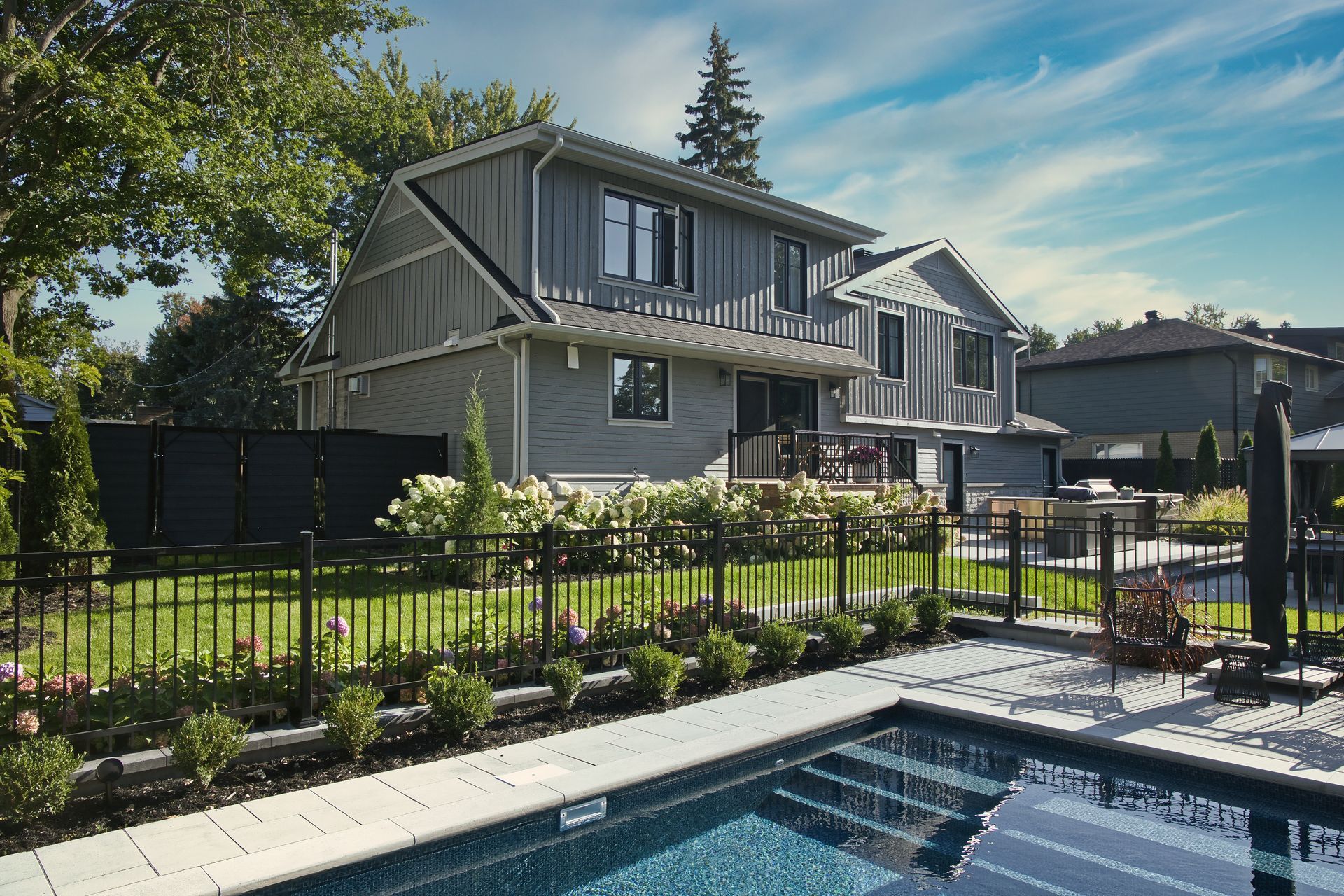 Modern two-story house with a backyard pool, green lawn, and black fencing on a sunny day.