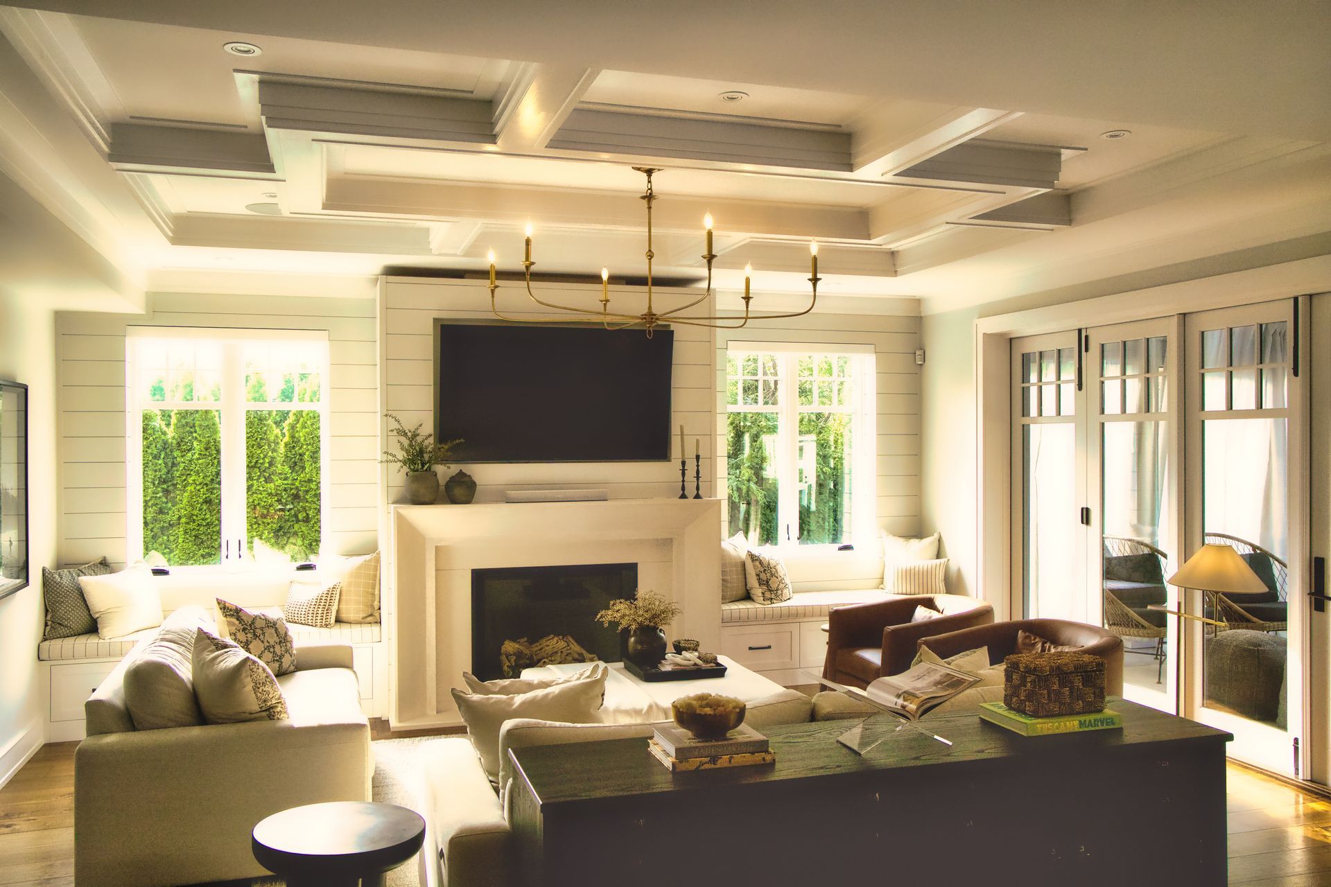 Living room with fireplace, white walls, and coffered ceiling. Windows, built-in seating, and a chandelier are visible.