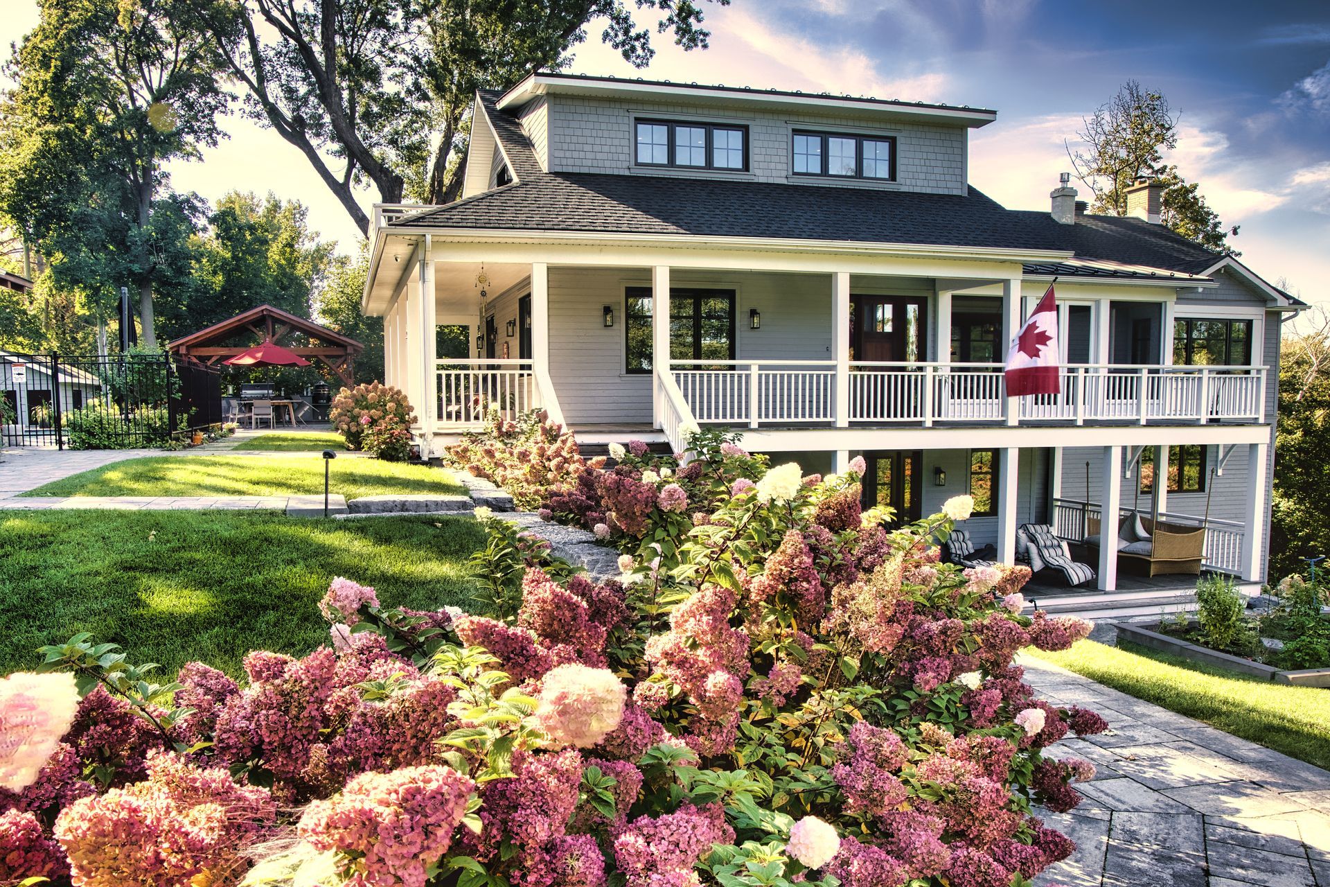 Two-story house with a porch and Canadian flag; pink hydrangeas in the foreground, sunny outdoor setting.