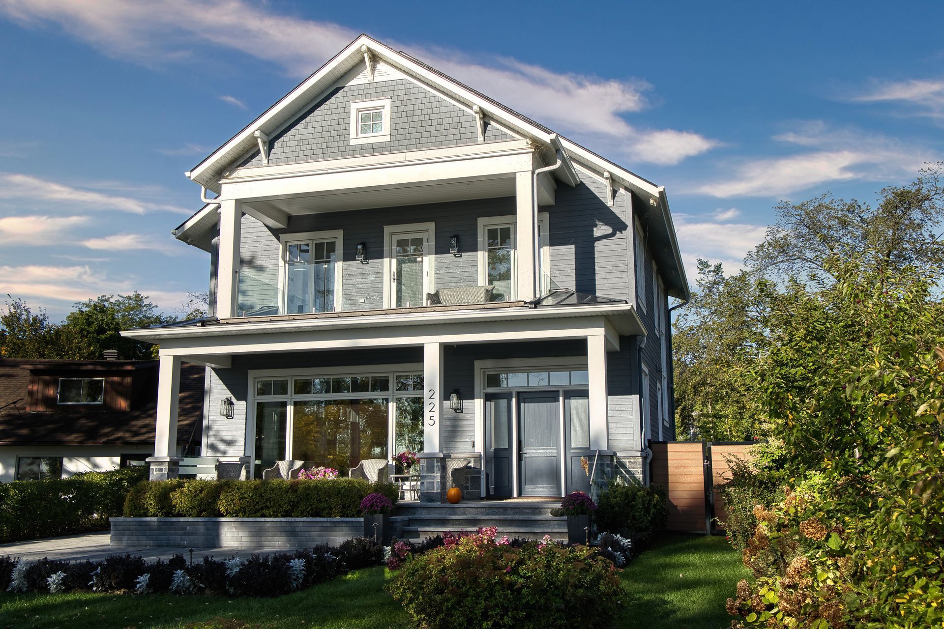 Two-story blue house with porch, balcony, and front yard landscaping on a sunny day.