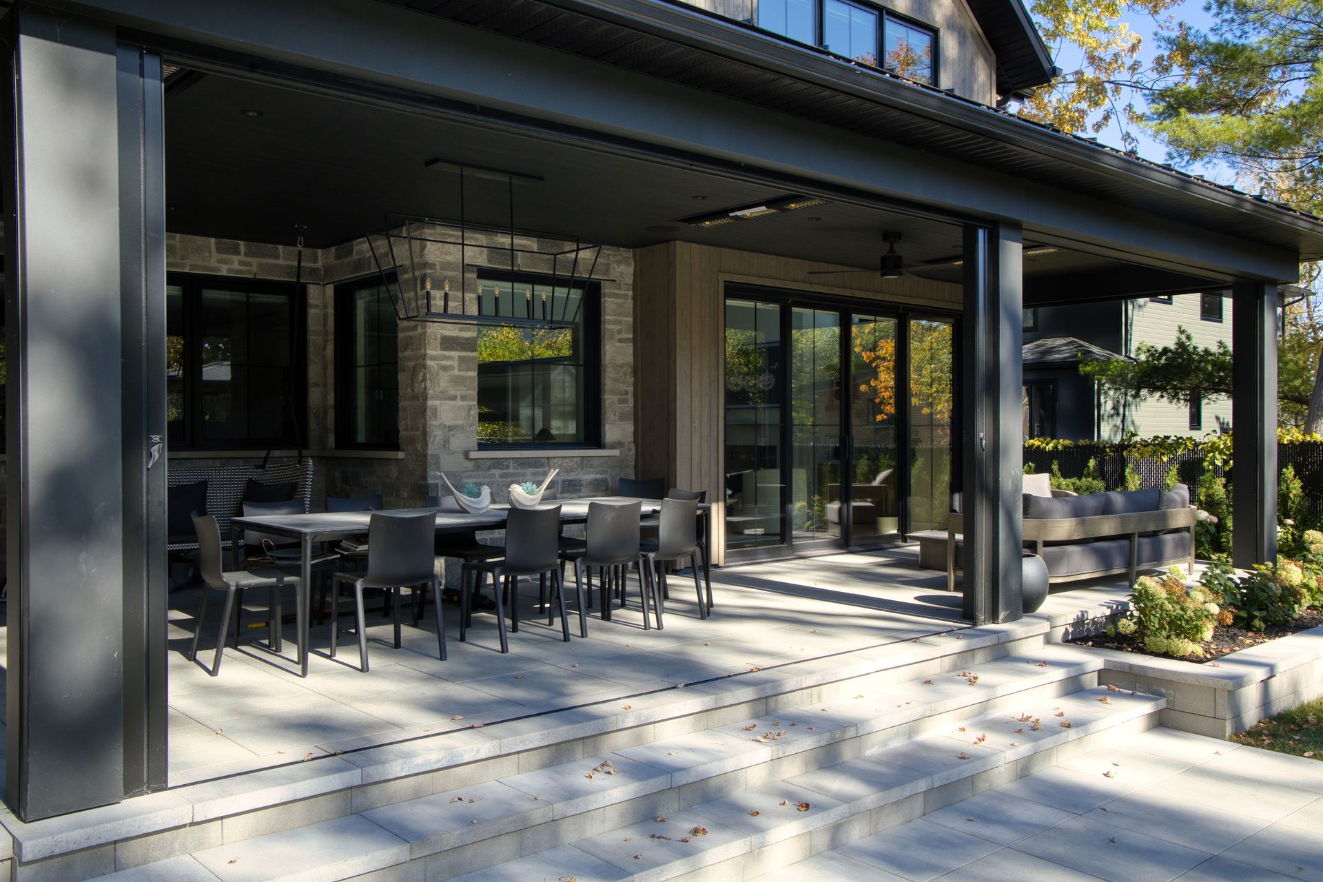 Outdoor patio with dining table and seating, under a black awning, adjacent to a stone and glass house.