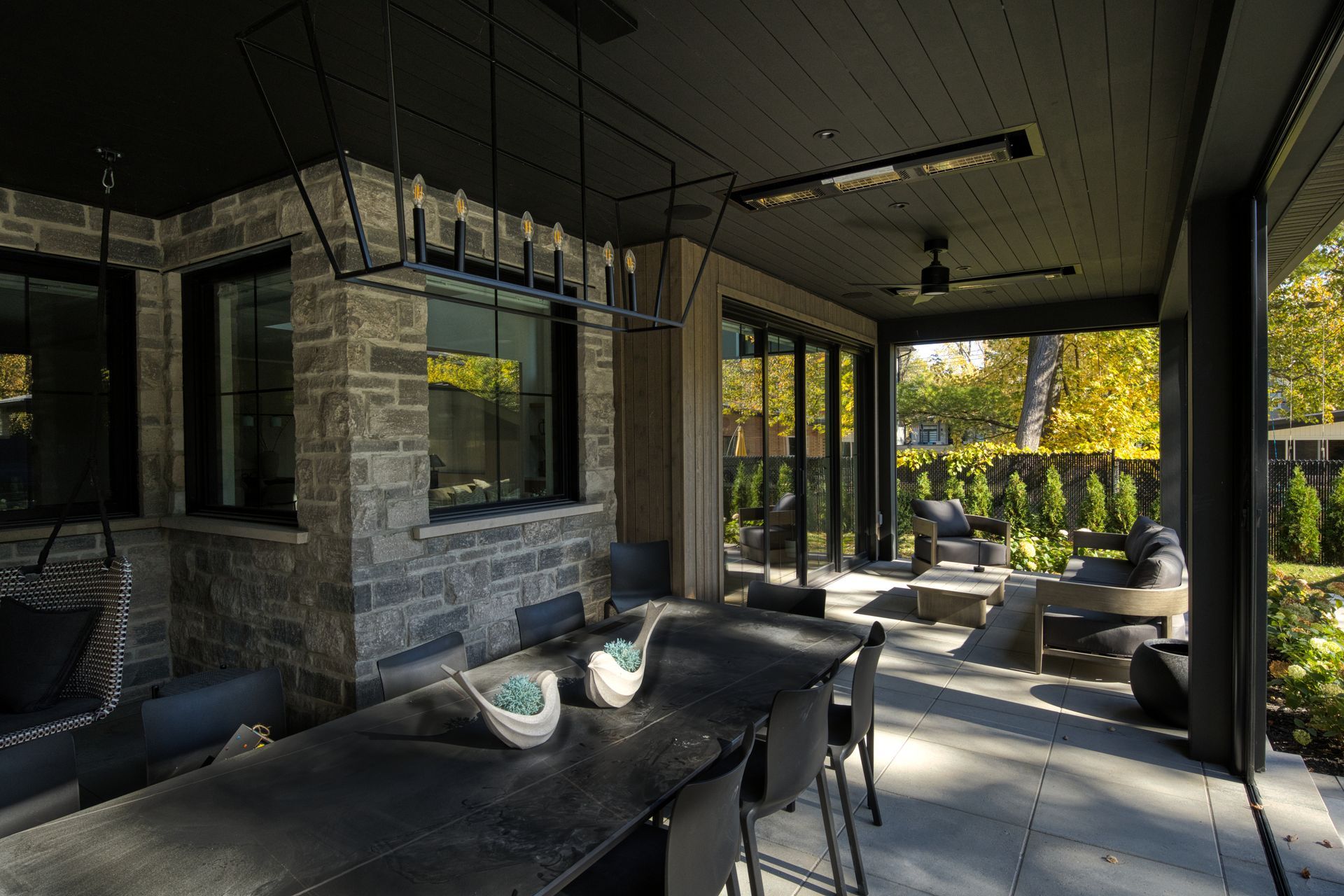 Covered outdoor dining area with dark gray ceiling, stone wall, and black table, chairs, and sliding glass doors to a backyard.