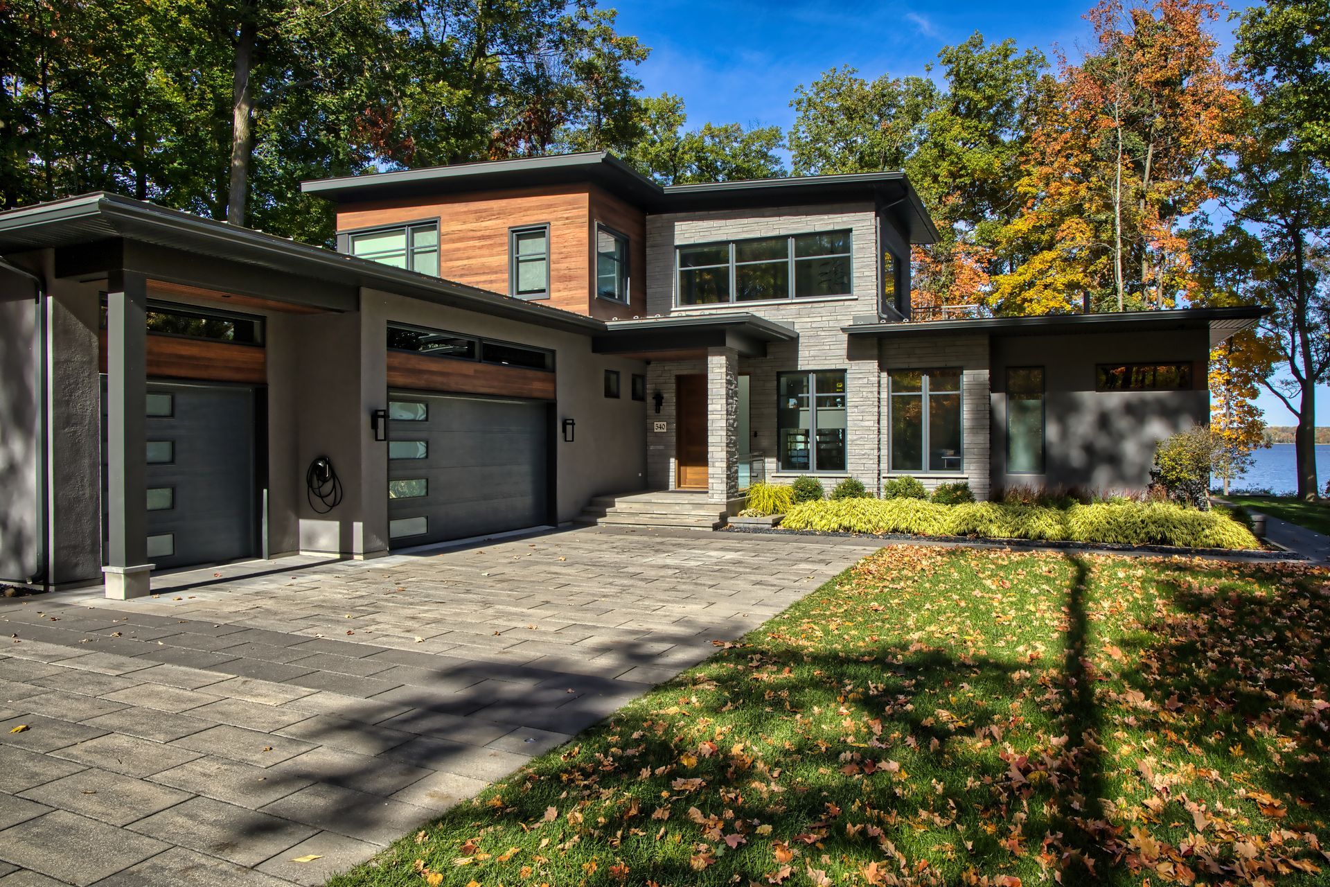 Modern two-story house with a gray facade, wooden accents, and a stone driveway, surrounded by trees.