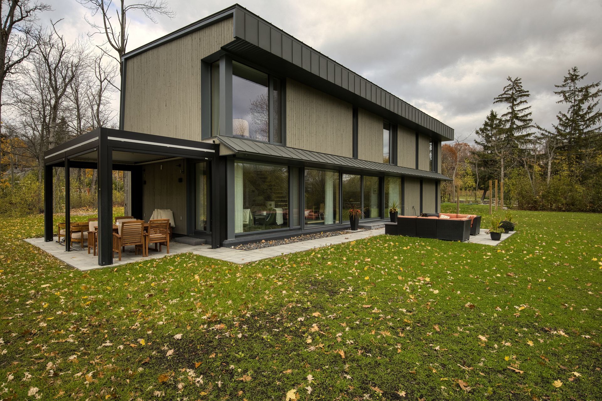 Modern two-story house with large windows, covered patio, and outdoor seating on a grassy lawn.