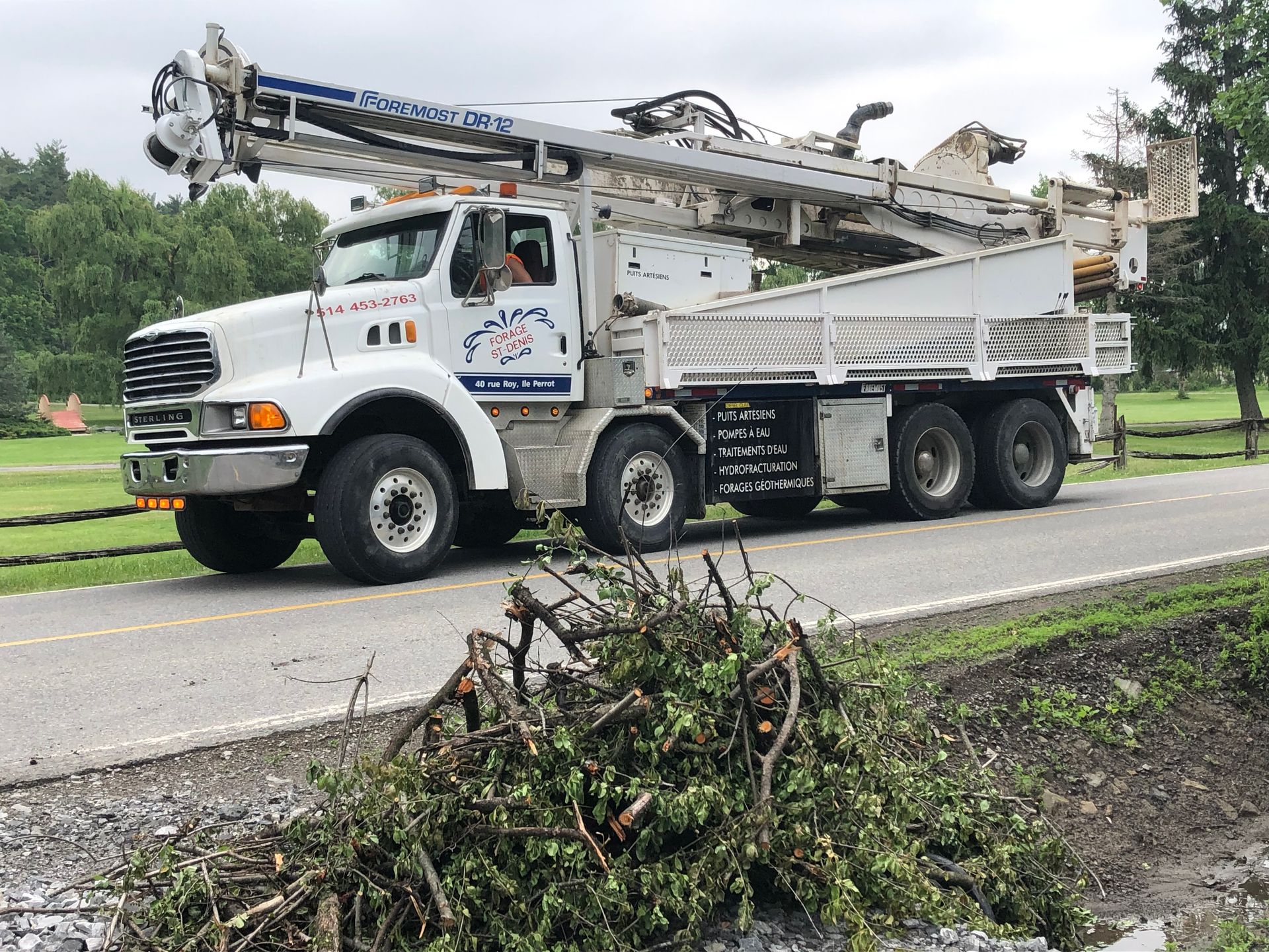 White truck with a drilling rig driving on a road next to a pile of brush.