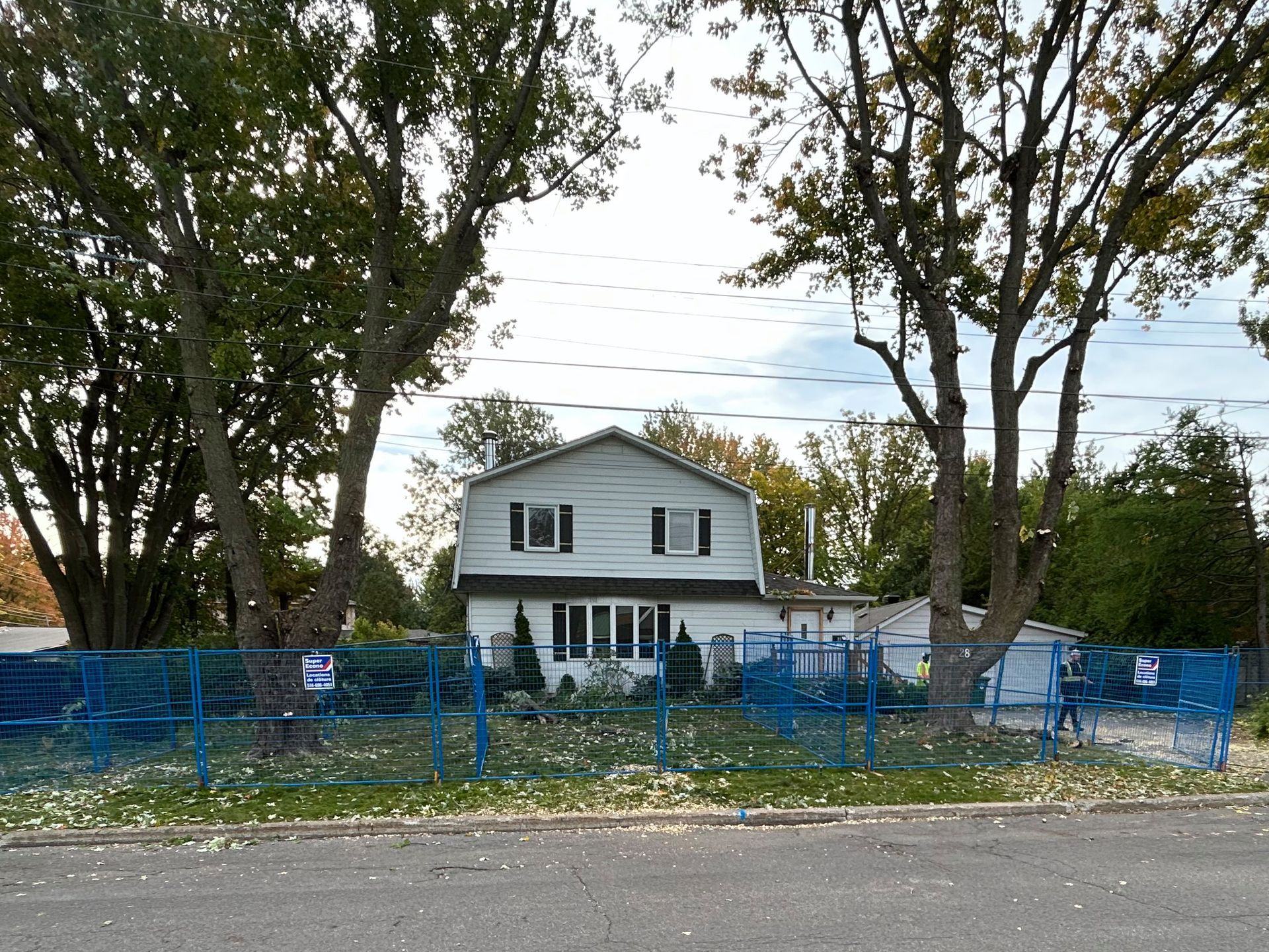 Two-story white house with blue fencing. Trees frame the house, leaves on the ground.