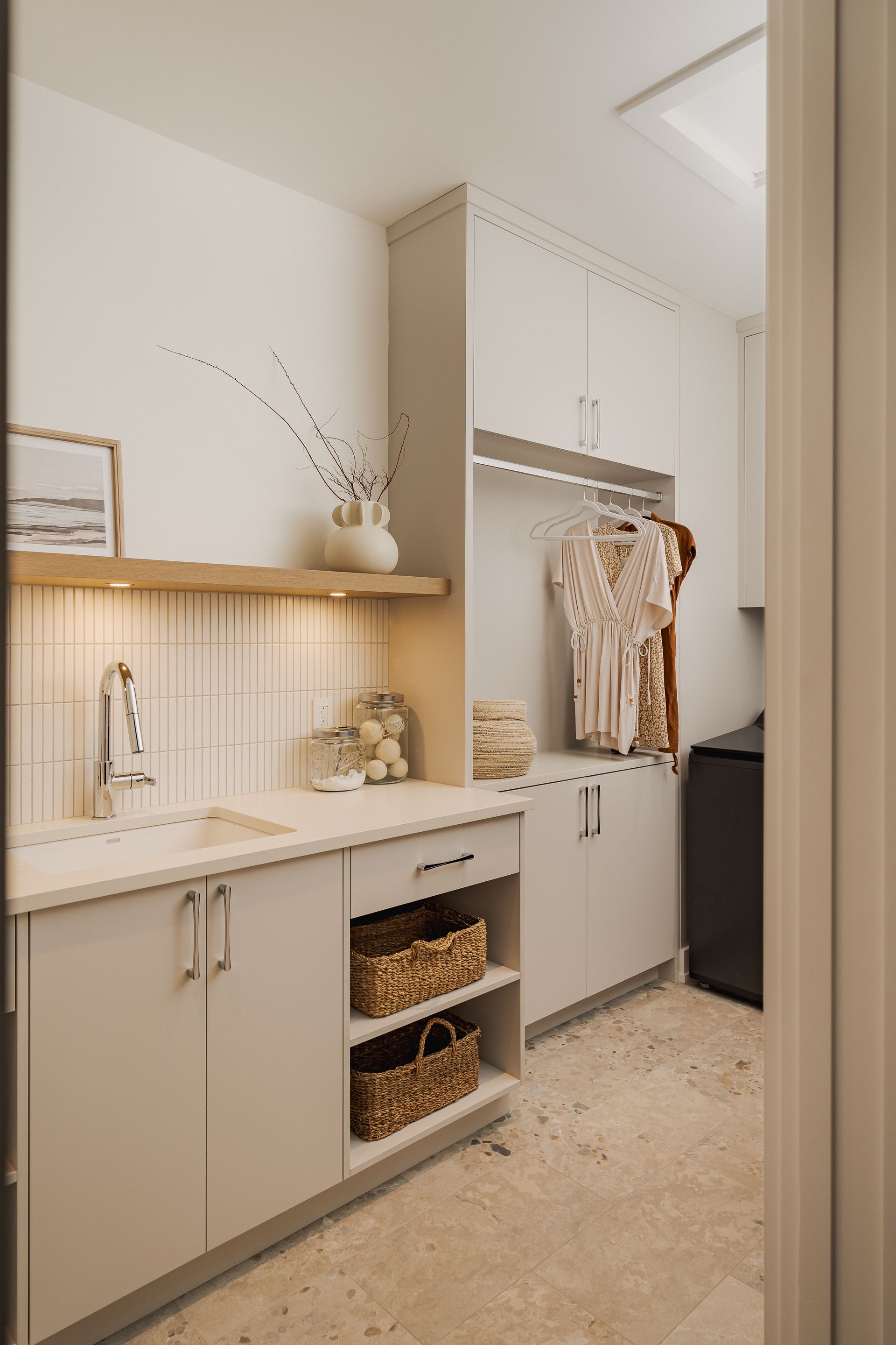 Laundry room with white cabinets, sink, open shelves, clothes hanging, and woven baskets.