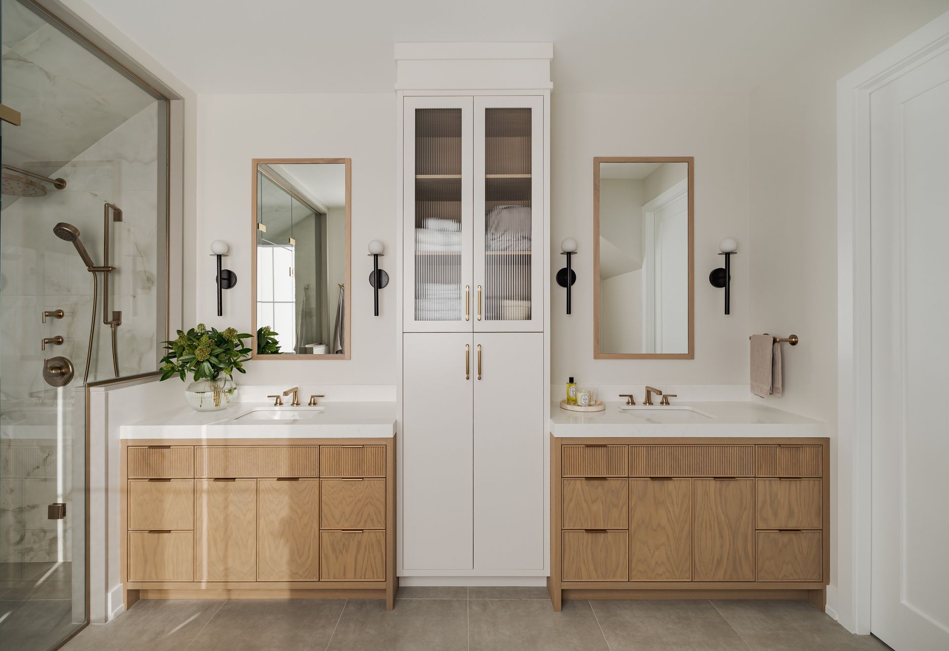 Bathroom with two wooden vanities, mirrors, and a tall cabinet; neutral tones.