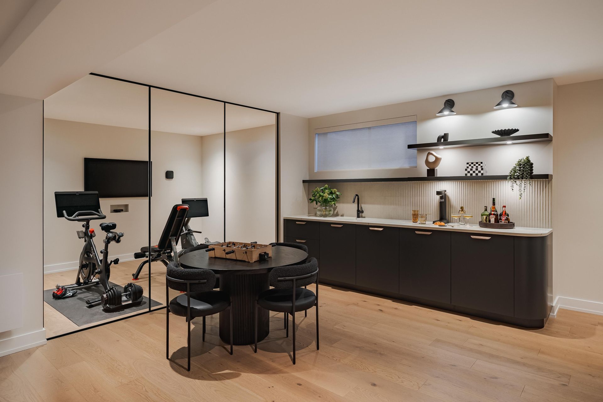 Modern basement with gym and bar. Black cabinets, shelves, round table, exercise bikes, mirrored wall.