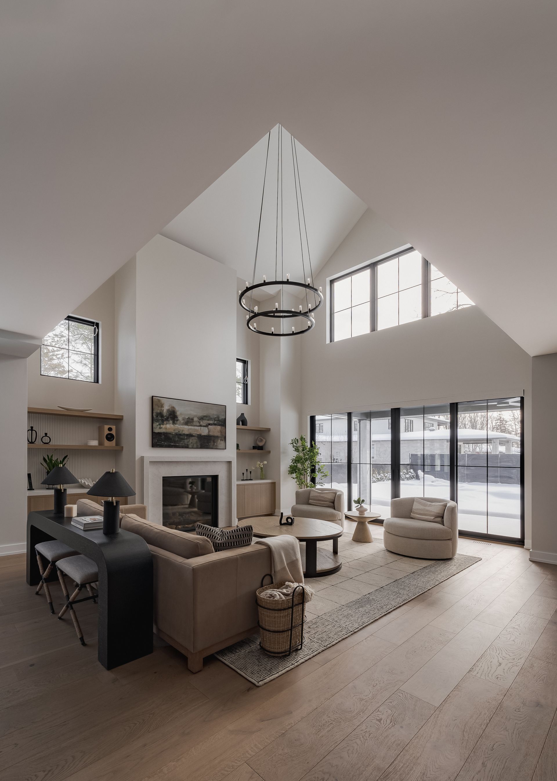 Bright, modern living room with high ceilings, fireplace, and large windows. Includes neutral-toned furniture and a statement chandelier.