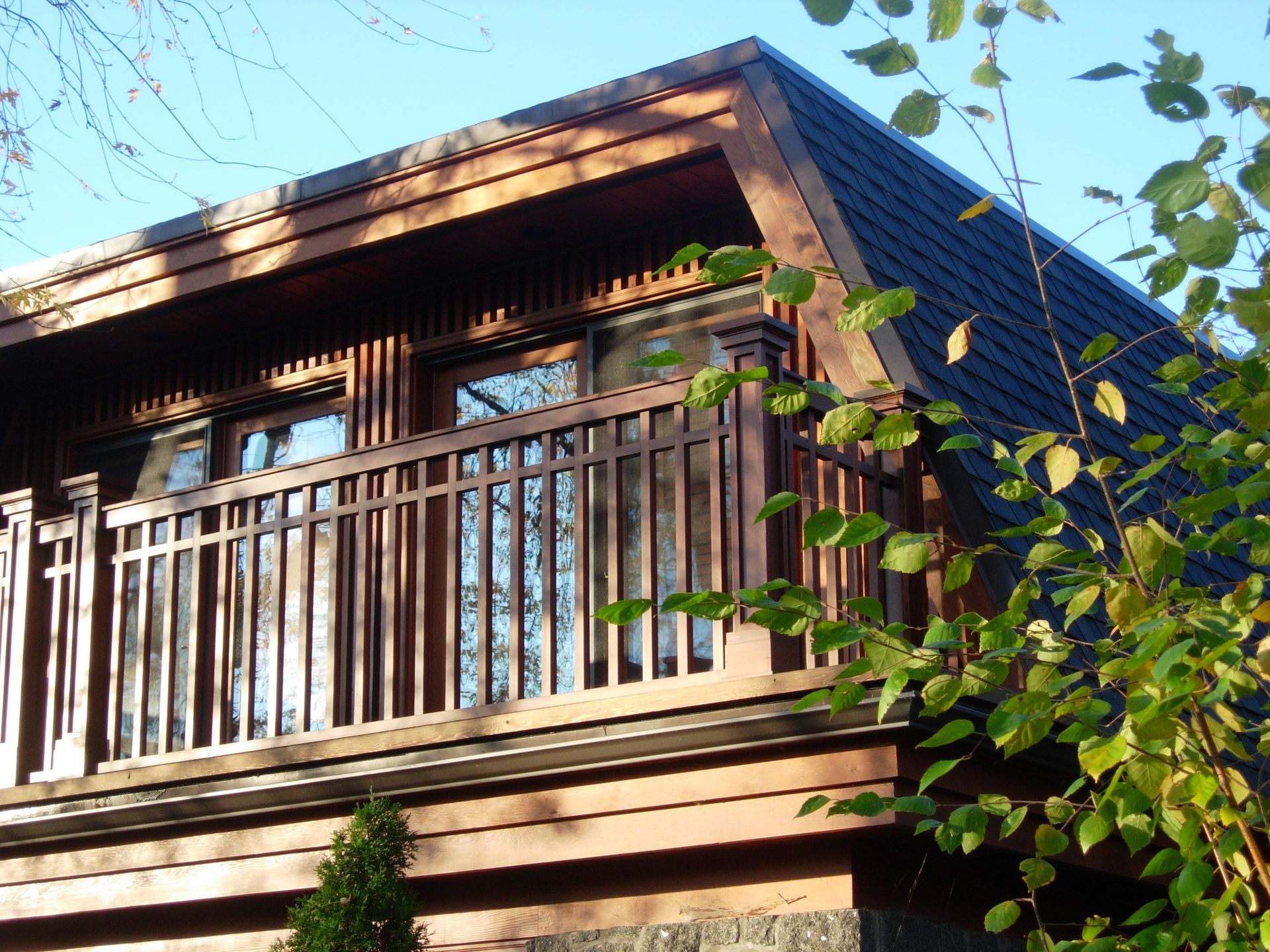 Brown wooden building with balcony, dark roof, against a blue sky, foliage in the foreground.
