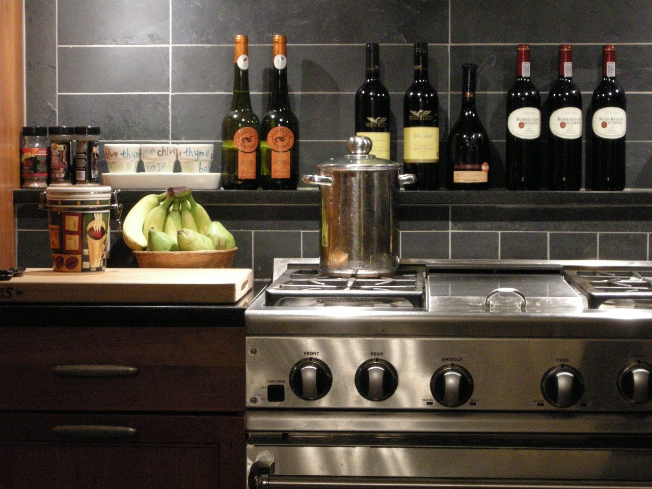 Stainless steel stove with a pot, bananas, and wine bottles in a kitchen.