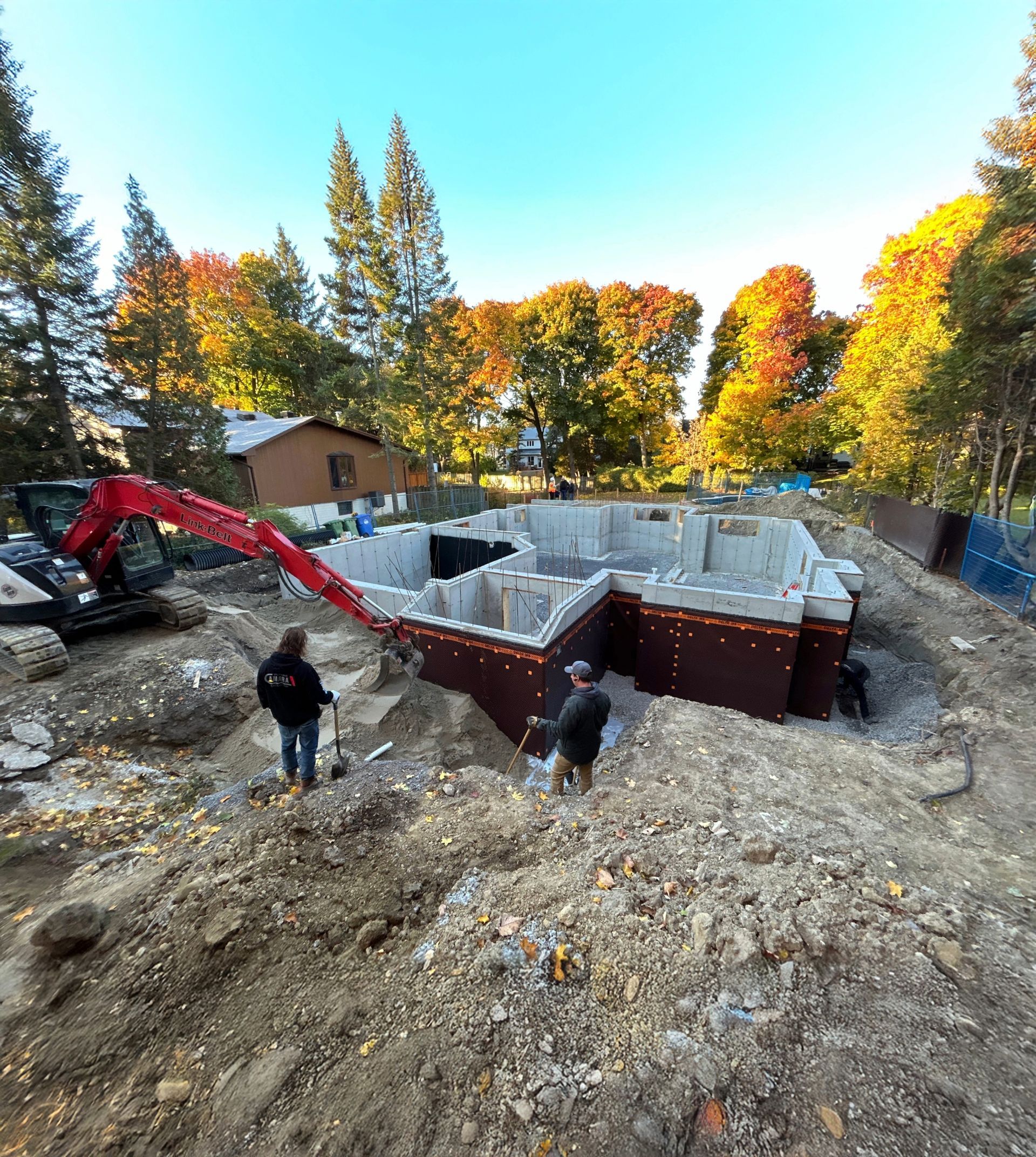 Construction site with foundation walls, workers, and a red excavator. Fall foliage in background.
