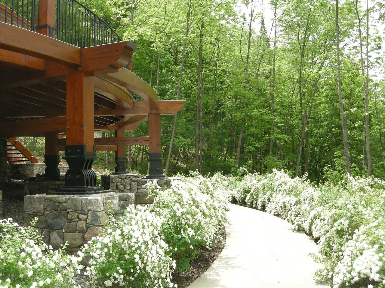 Wooden deck with stone pillars overlooking a path lined with white flowering bushes, leading into a forest.