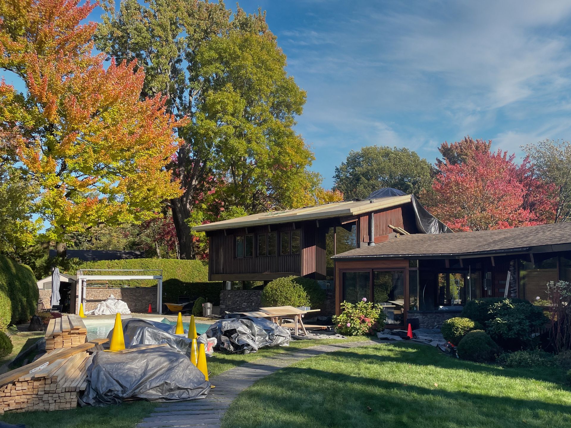 House with brown exterior and large trees with fall foliage. Construction materials in the foreground.