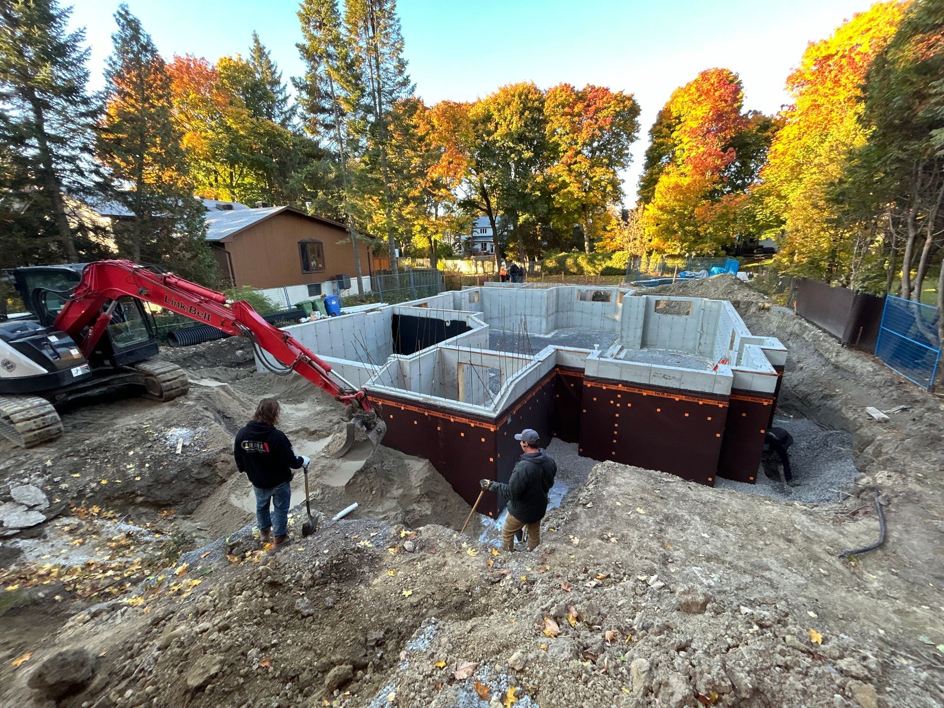 Construction site with workers, excavator, and foundation walls wrapped in brown material, surrounded by fall foliage.