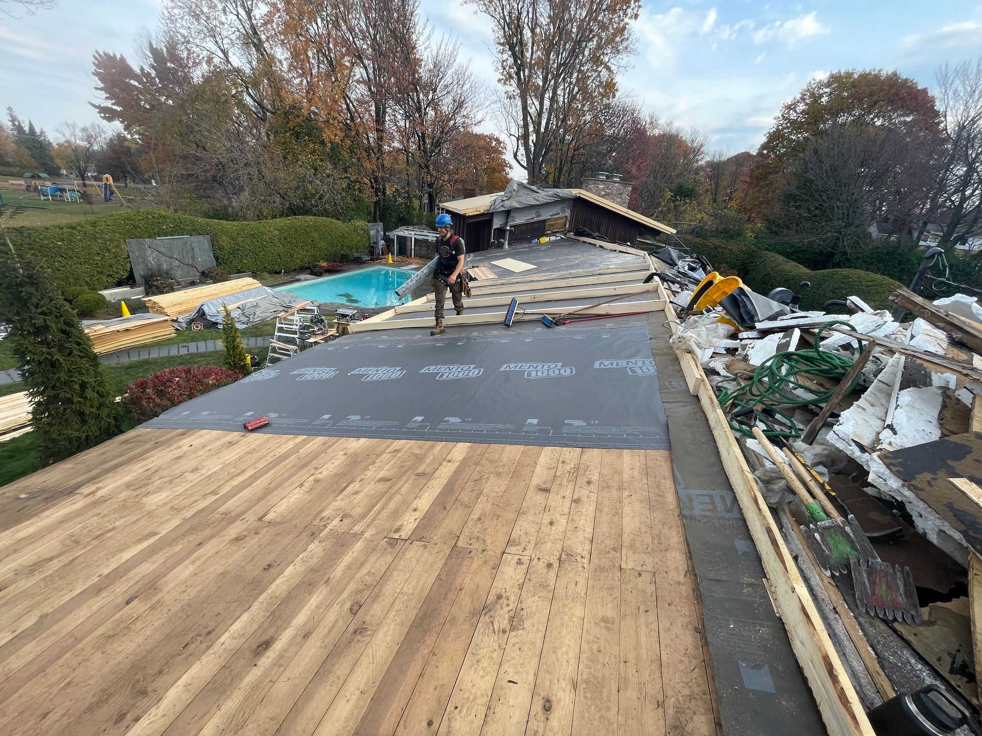 Roofer working on a flat roof, debris nearby, with a pool and trees in the background. Sunny day.