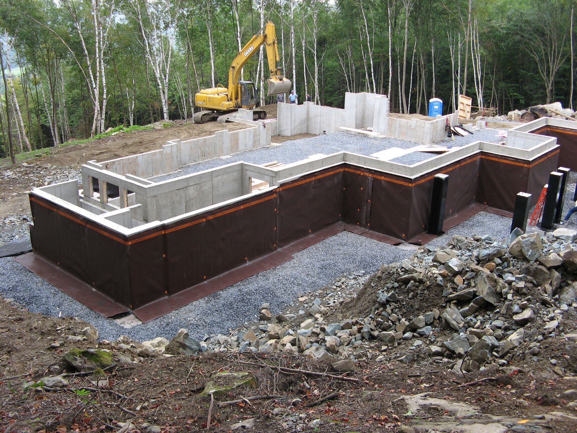 Foundation of a house under construction; a brown membrane covers the concrete walls with an excavator in the background.