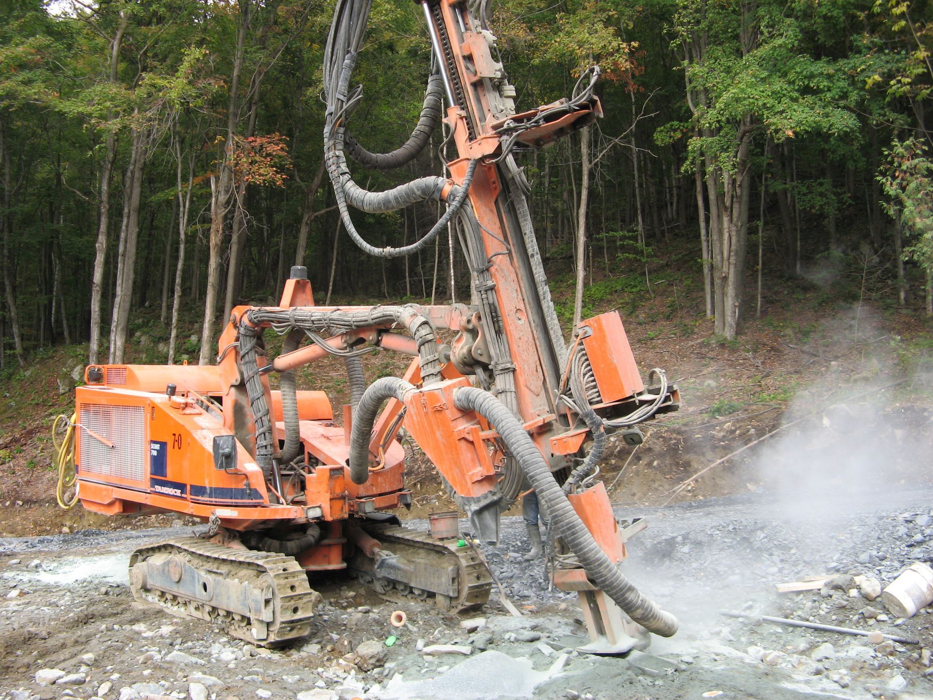 Orange drill rig boring into rock; trees in the background.