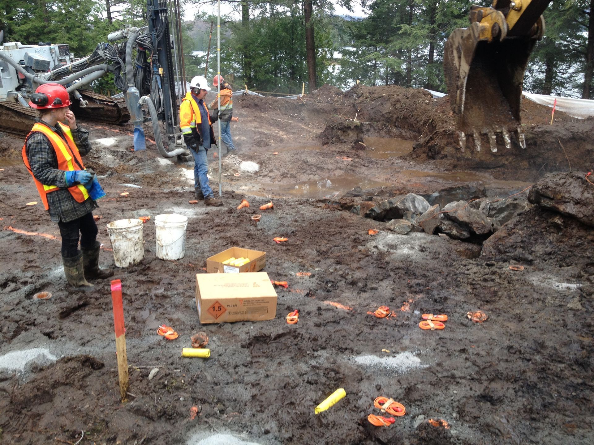 Construction site. Workers in safety gear, muddy ground with equipment and boxes. Excavator in background.